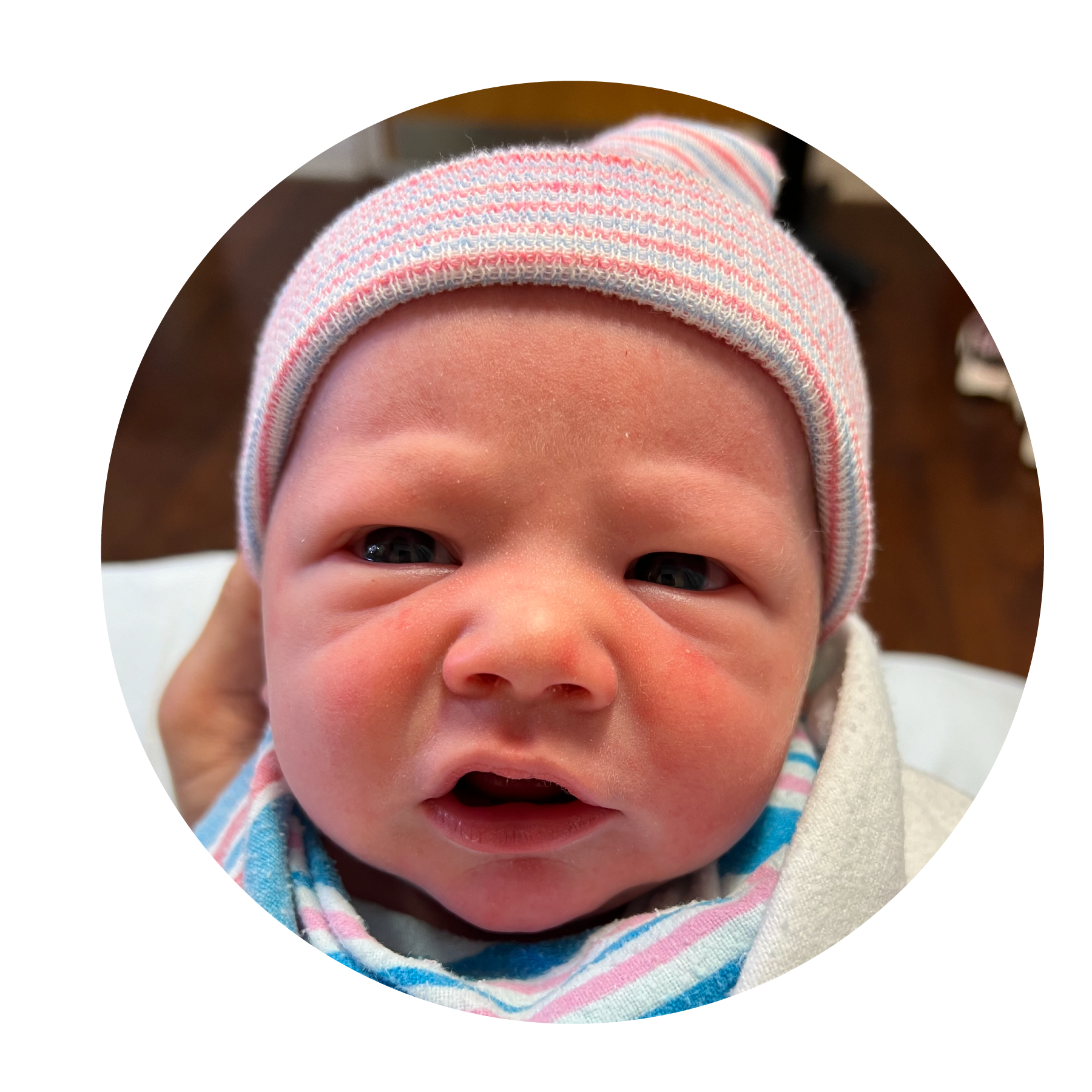 Close-up of a newborn baby wearing a striped hat and blanket, with a slightly open mouth and curious eyes.