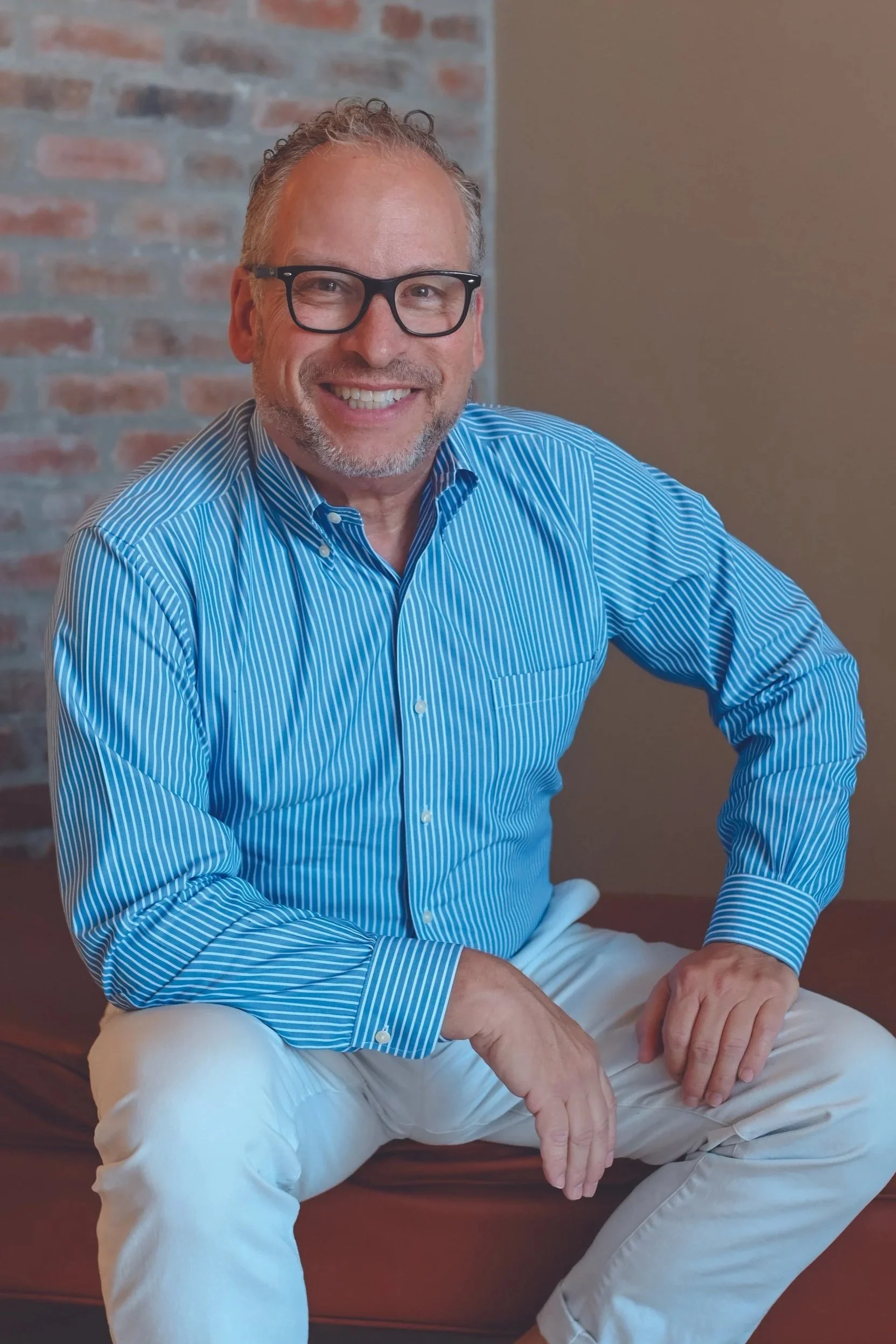 A smiling middle-aged man with glasses and gray hair, wearing a black sweater over a light blue collared shirt, standing outdoors near water with rocky terrain in the background.