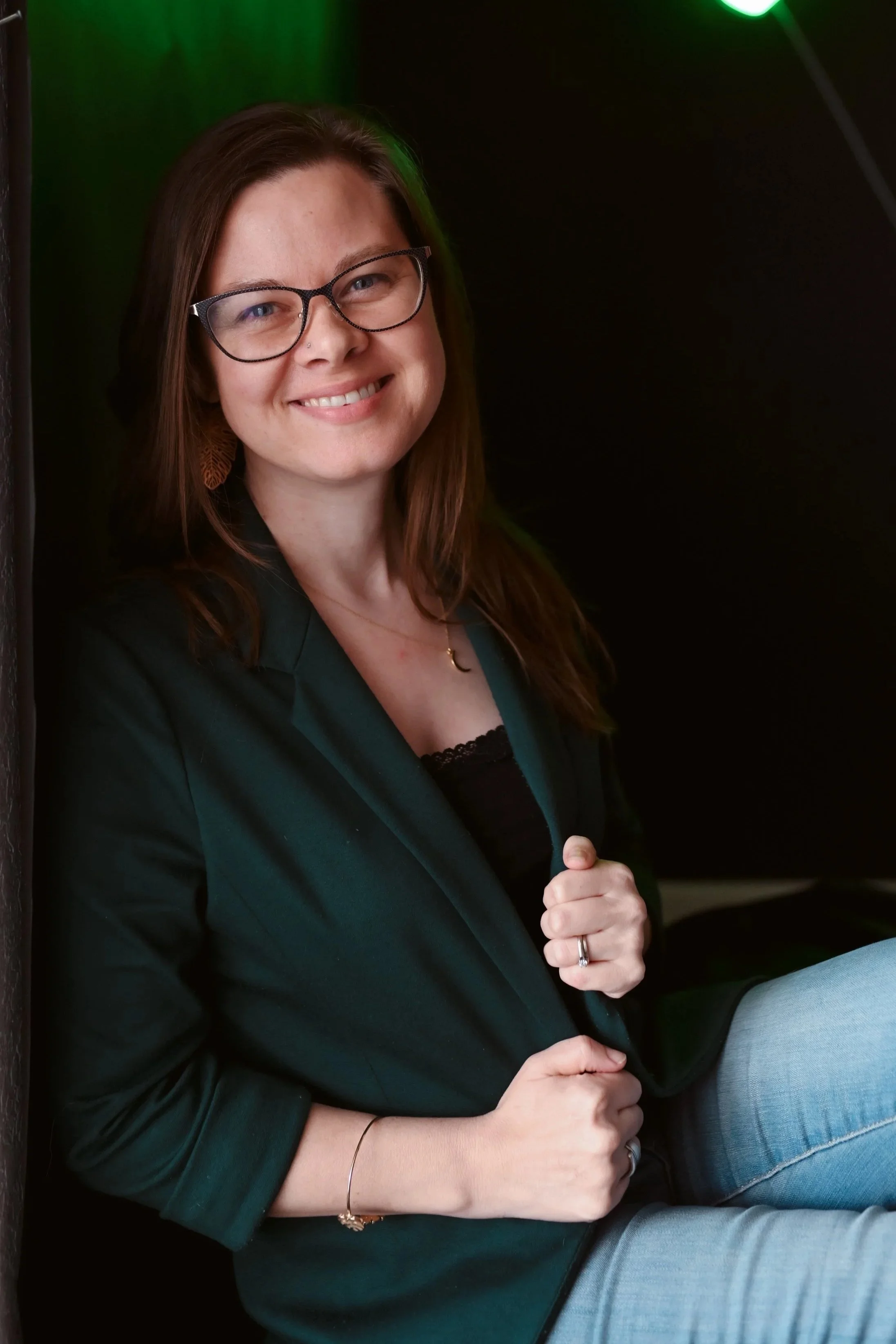 A woman with long brown hair, glasses, and a black shirt smiling outdoors near a waterfall.