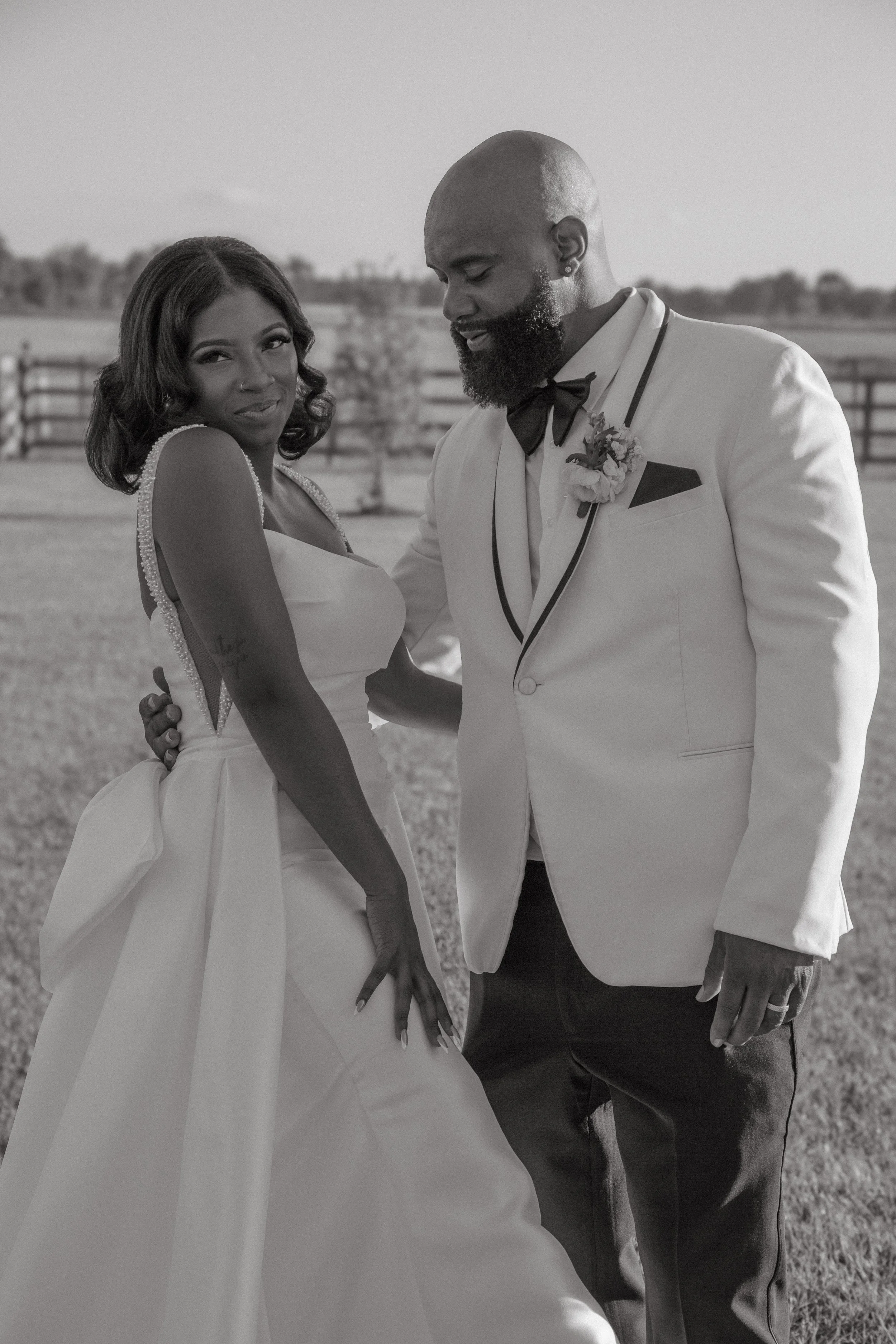 A black-and-white photo of a couple dressed in wedding attire outdoors. The woman is wearing a strapless wedding gown with pearl details on the straps, and the man is in a tuxedo with a bow tie and boutonniere. They are standing close together, holding hands, with a fence and open landscape in the background.