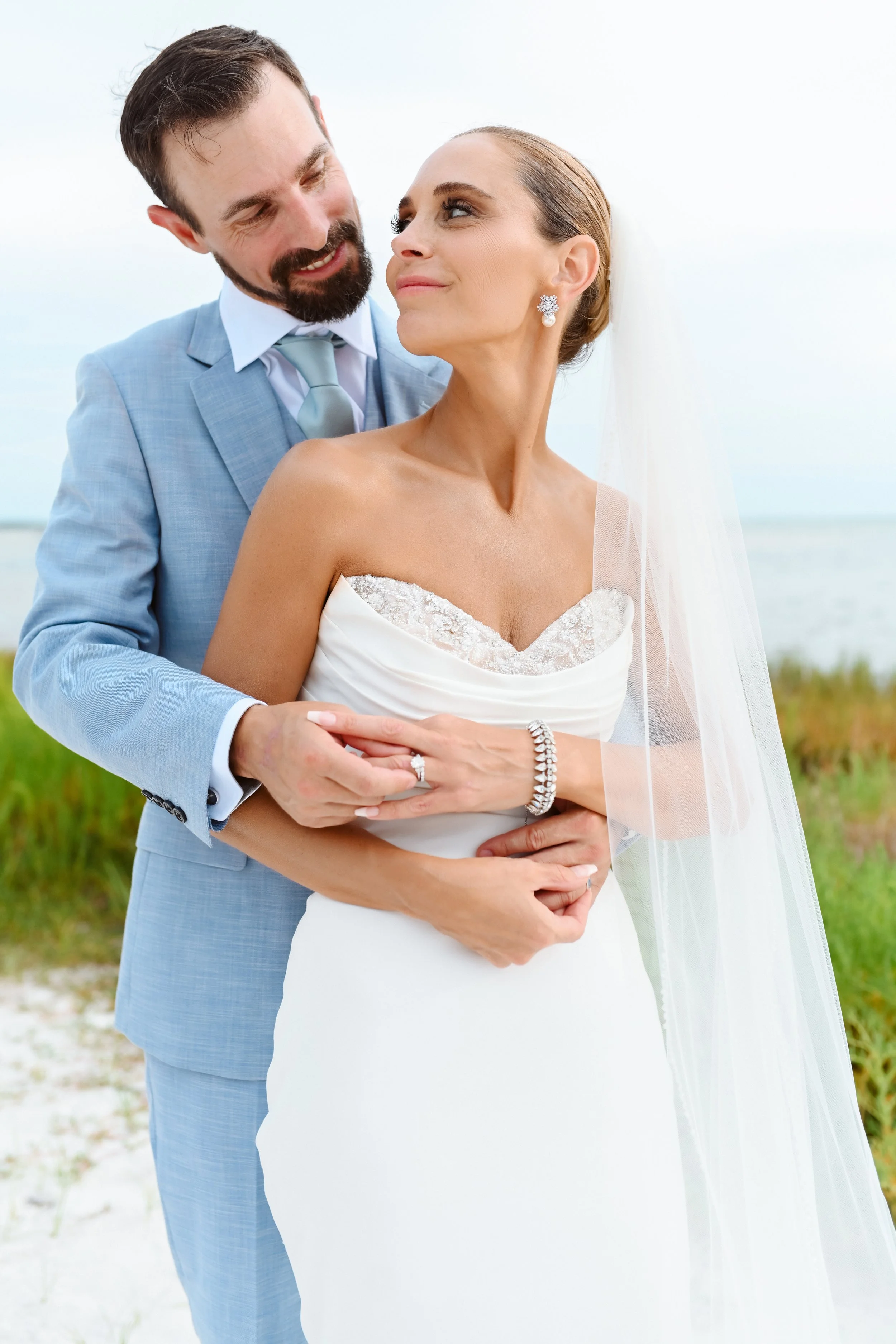 A bride and groom embracing on a beach, looking into each other's eyes.