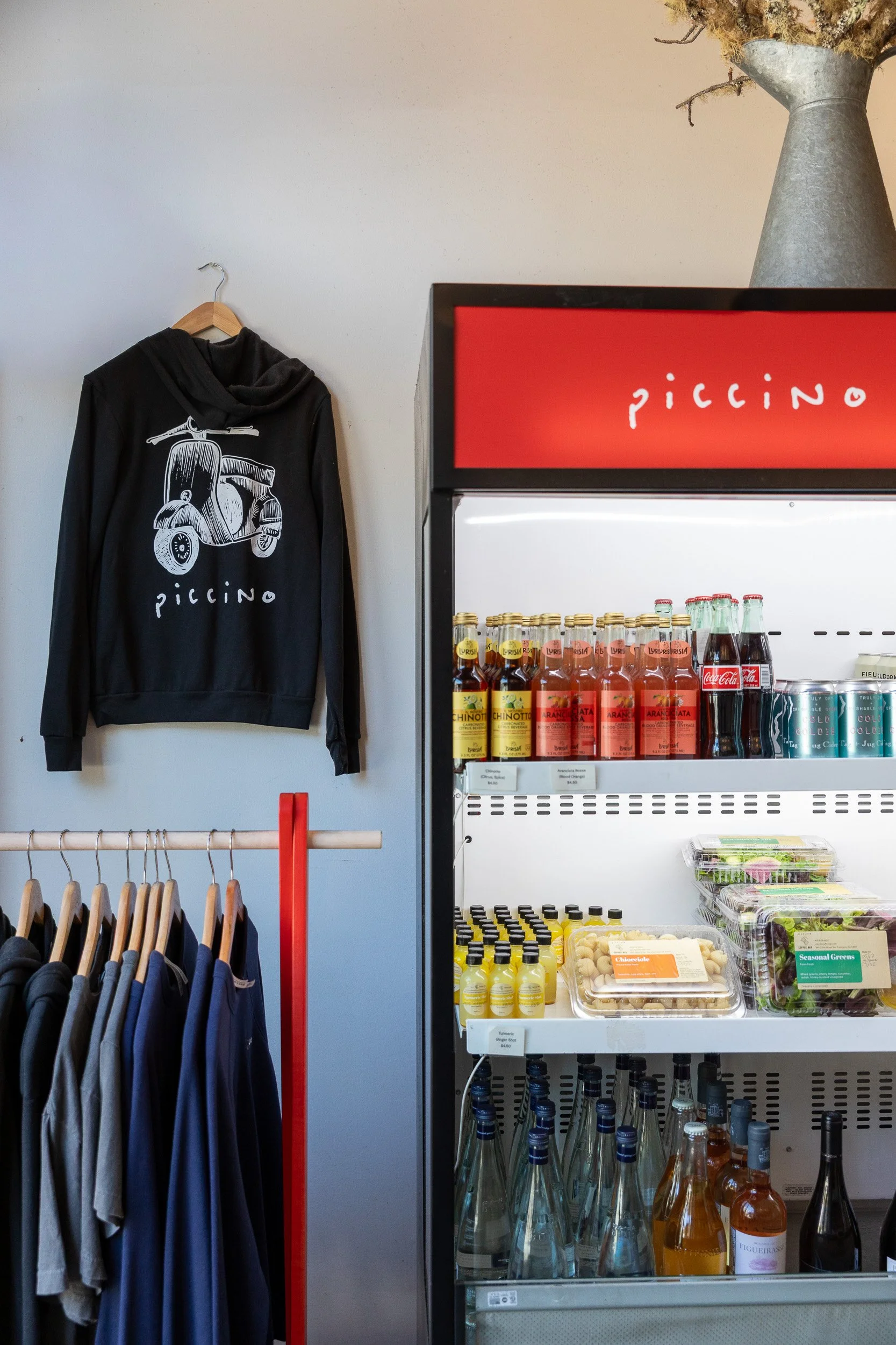 Piccino swag: branded Piccino sweatshirts and crew necks next to a take-out fridge with fresh pasta, prepared sandwiches, beer, and wine