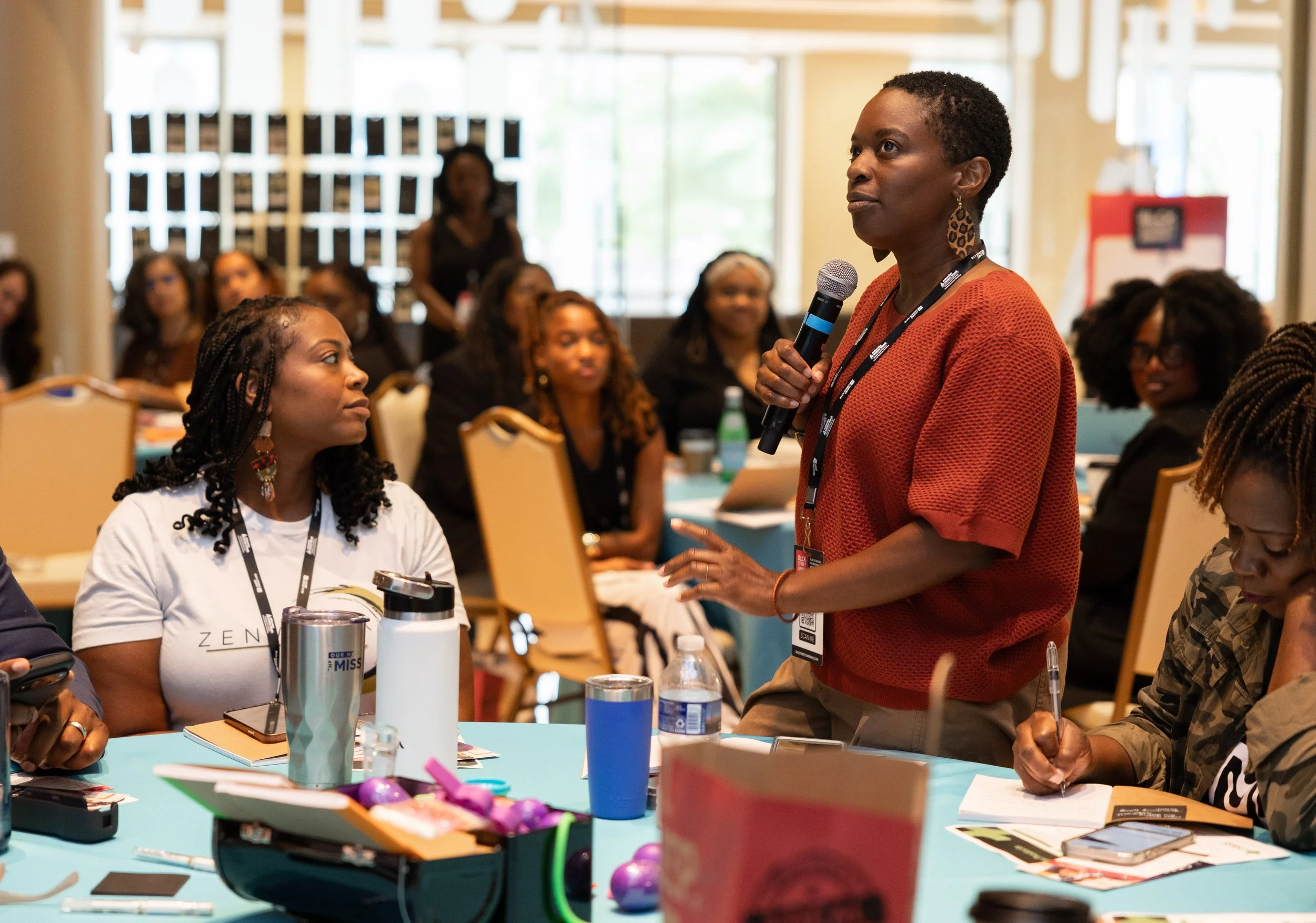 An attendee stands to speak with a microphone during a BLCK Street Summit 2025 session, as other participants listen from tables in the conference room.