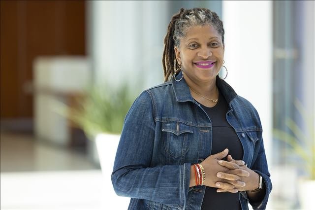 A woman with curly hair in dreadlocks, wearing a denim jacket, black shirt, and jewelry, smiling with hands clasped.