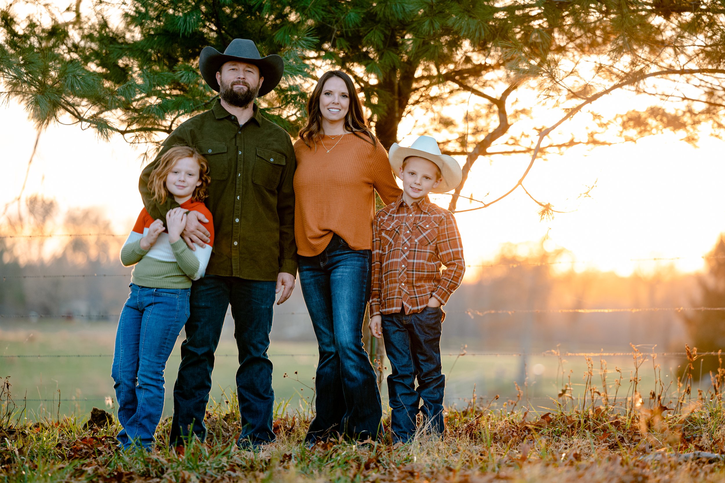 Family of four standing outdoors during sunset, with tall trees in the background. The father and son are wearing cowboy hats and plaid shirts, while the mother and daughter are dressed casually, all smiling.