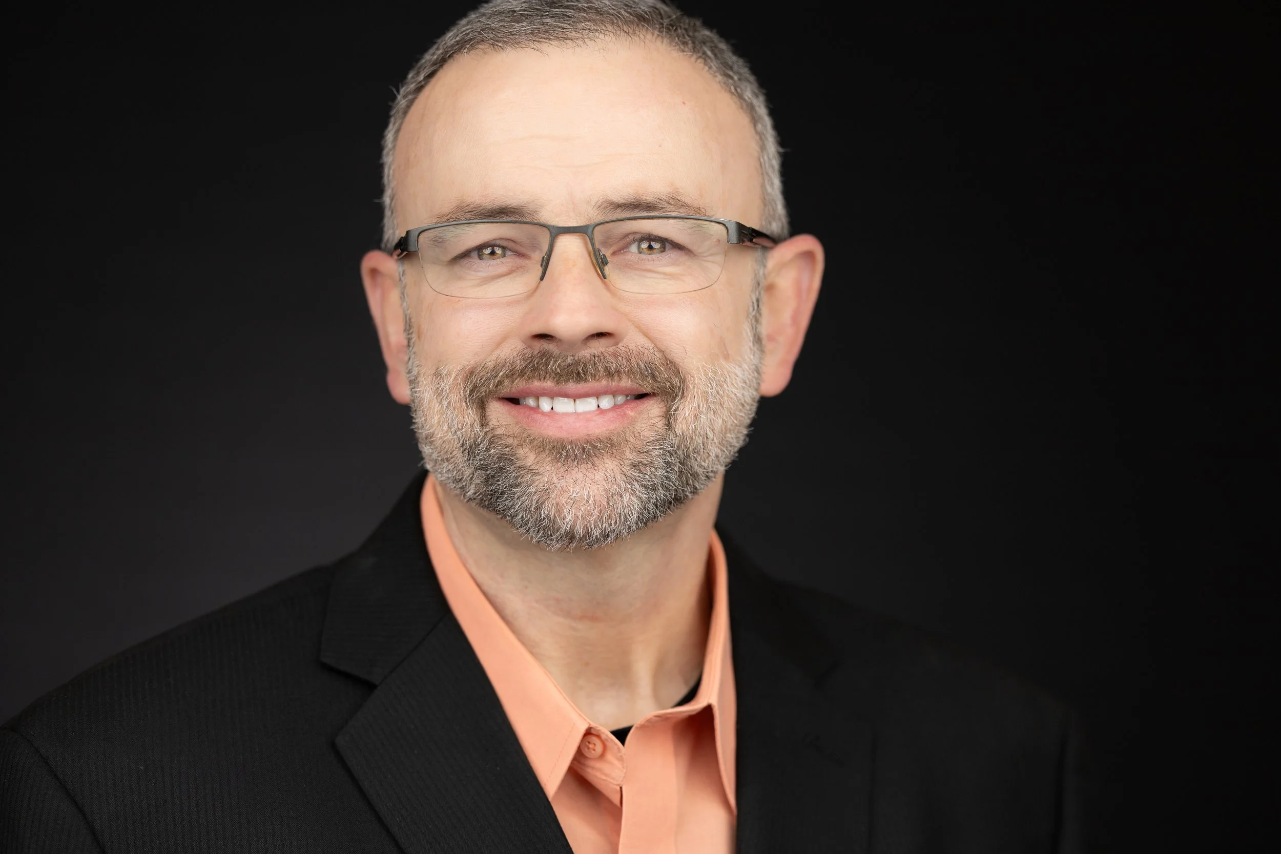 Headshot of a middle-aged man with short gray hair, glasses, a beard, and a friendly smile, wearing a black suit jacket and a peach-colored collared shirt, against a dark background.