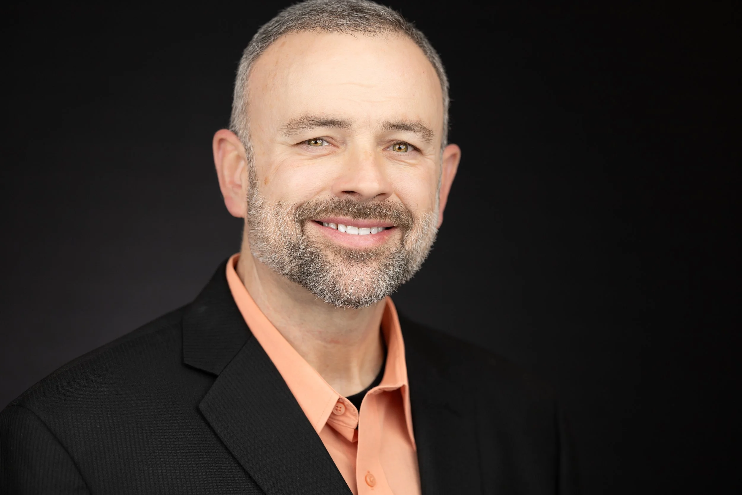 Headshot of a smiling middle-aged man with short gray hair, beard, green eyes, wearing a black blazer and peach-colored shirt against a black background.