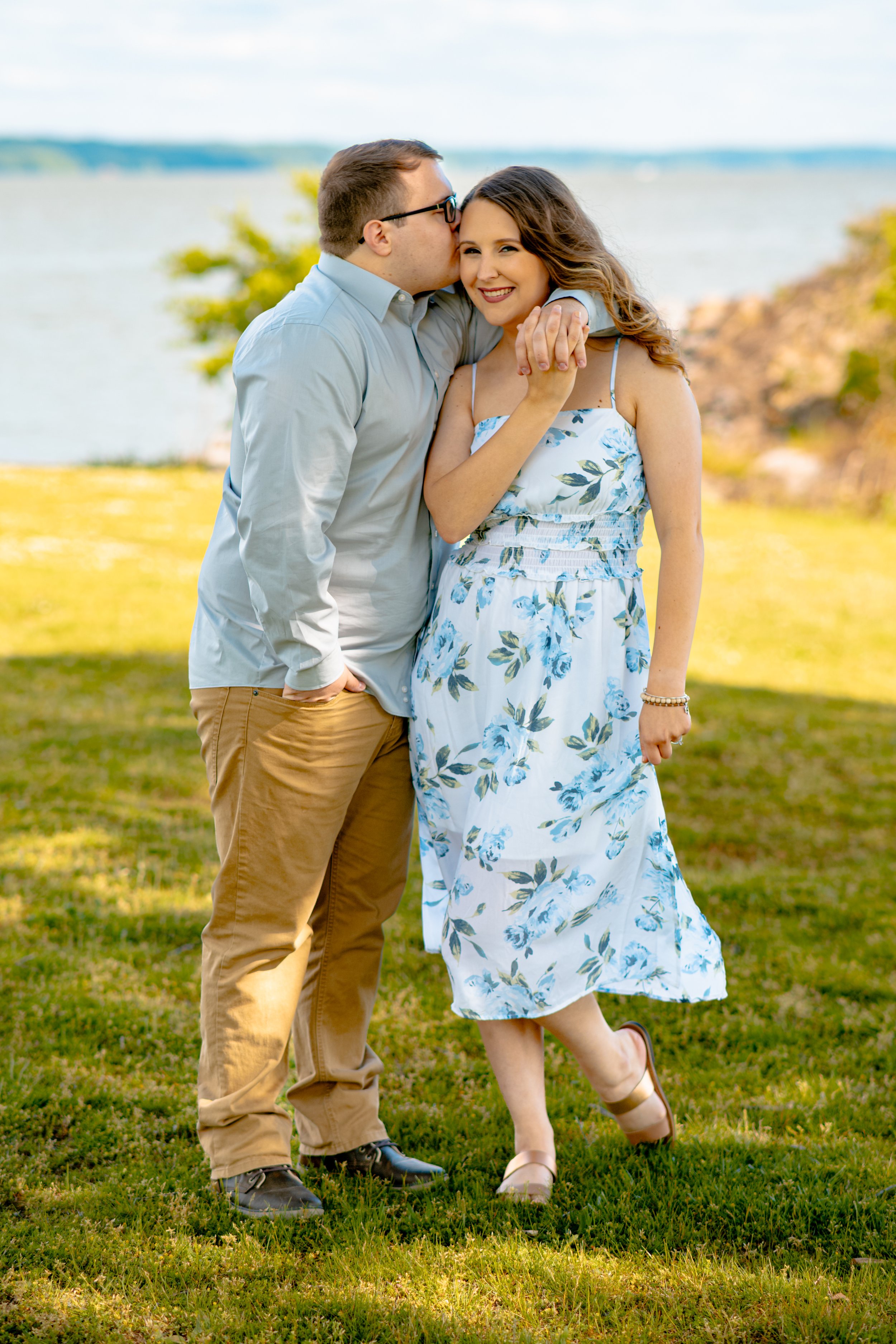 A man kisses a woman on the cheek while they smile, standing on grass near a body of water with trees and rocks in the background.