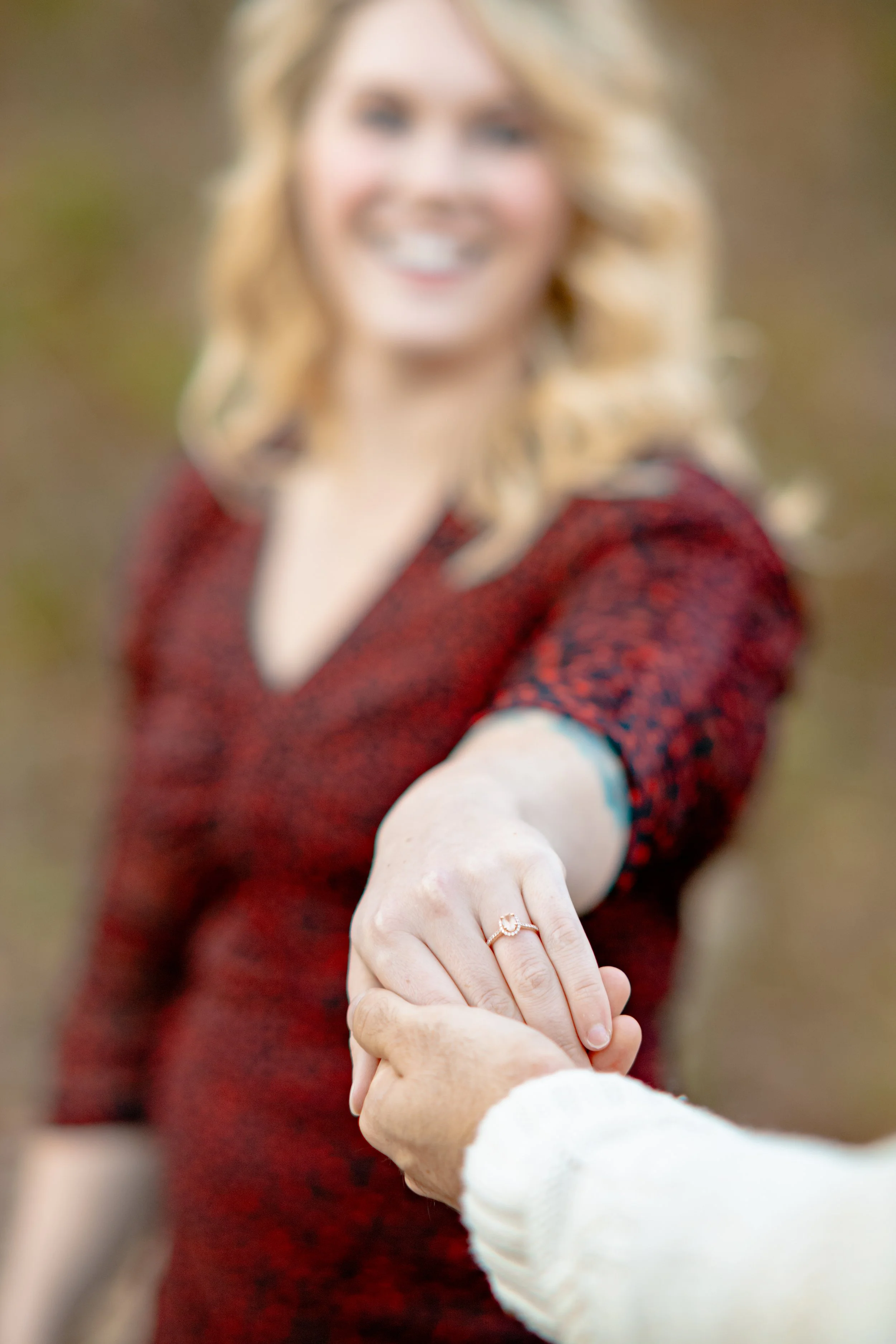 A woman with blonde, curly hair smiling and extending her hand, showing an engagement ring, in an outdoor setting.