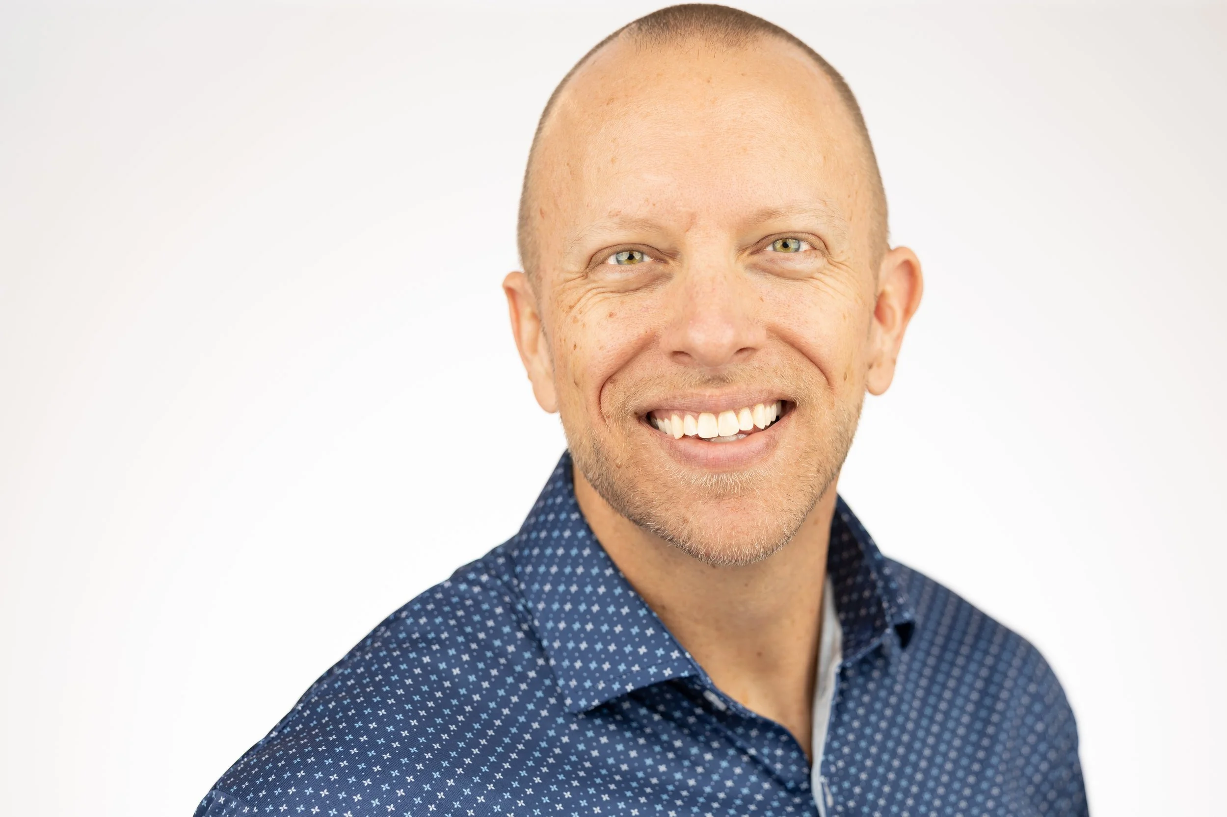 Smiling man with a bald head and light skin wearing a navy blue patterned shirt