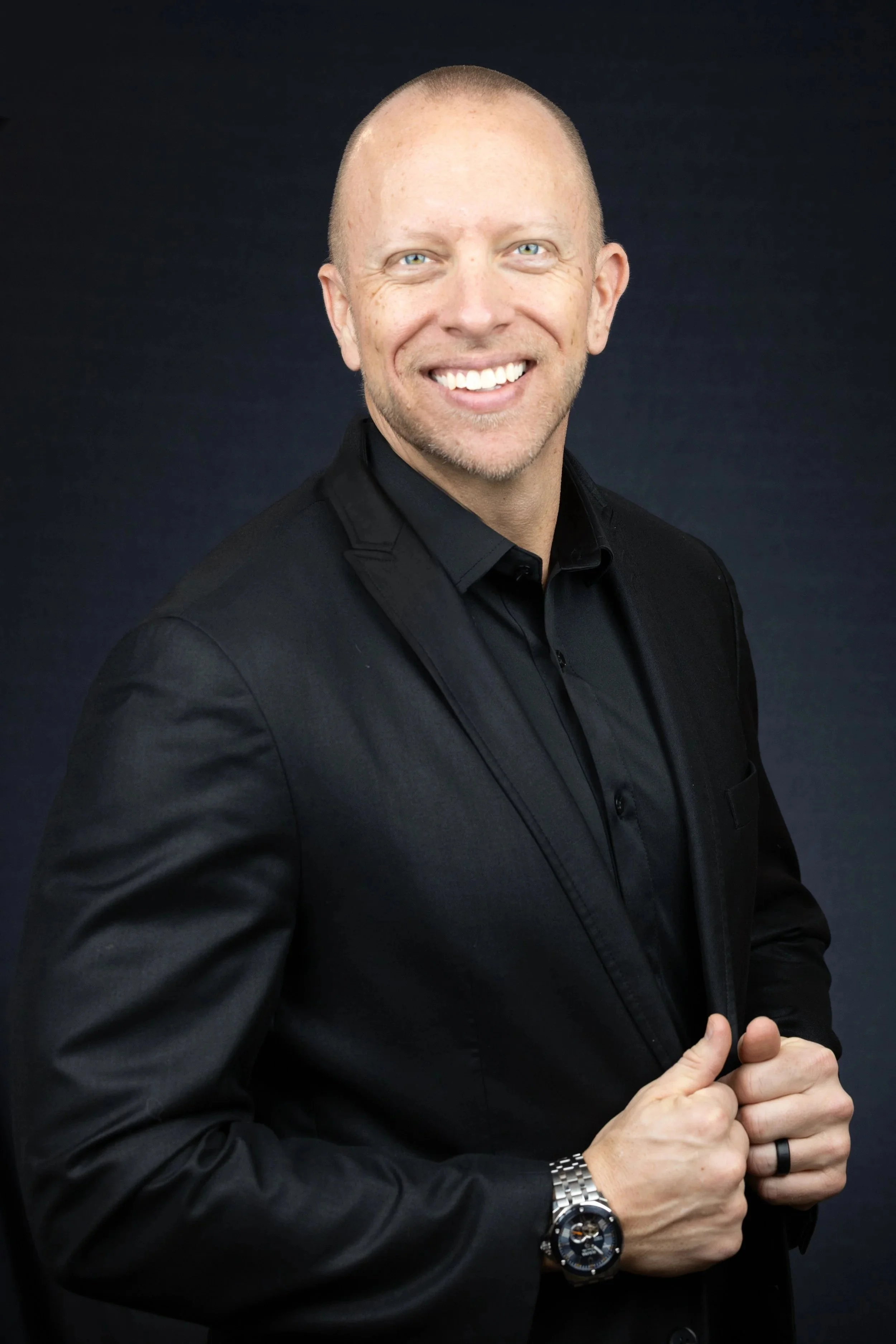 A portrait of a smiling man with a bald head, light skin, blue eyes, in a black suit and black shirt, wearing a silver watch and black ring, against a dark background.