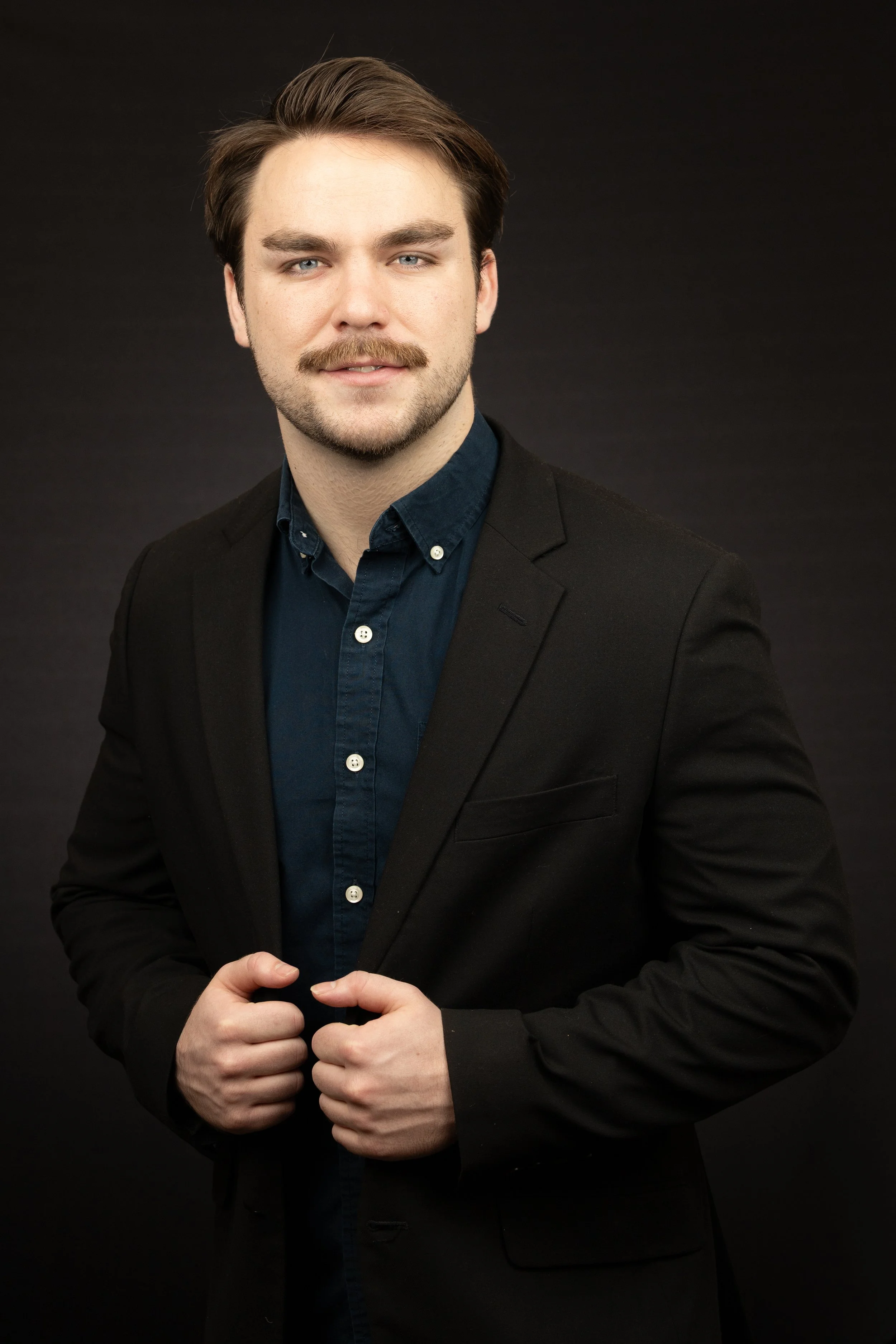 A young man with light skin, brown hair, and a mustache, wearing a black blazer and a dark blue button-up shirt, adjusting his blazer against a black background.