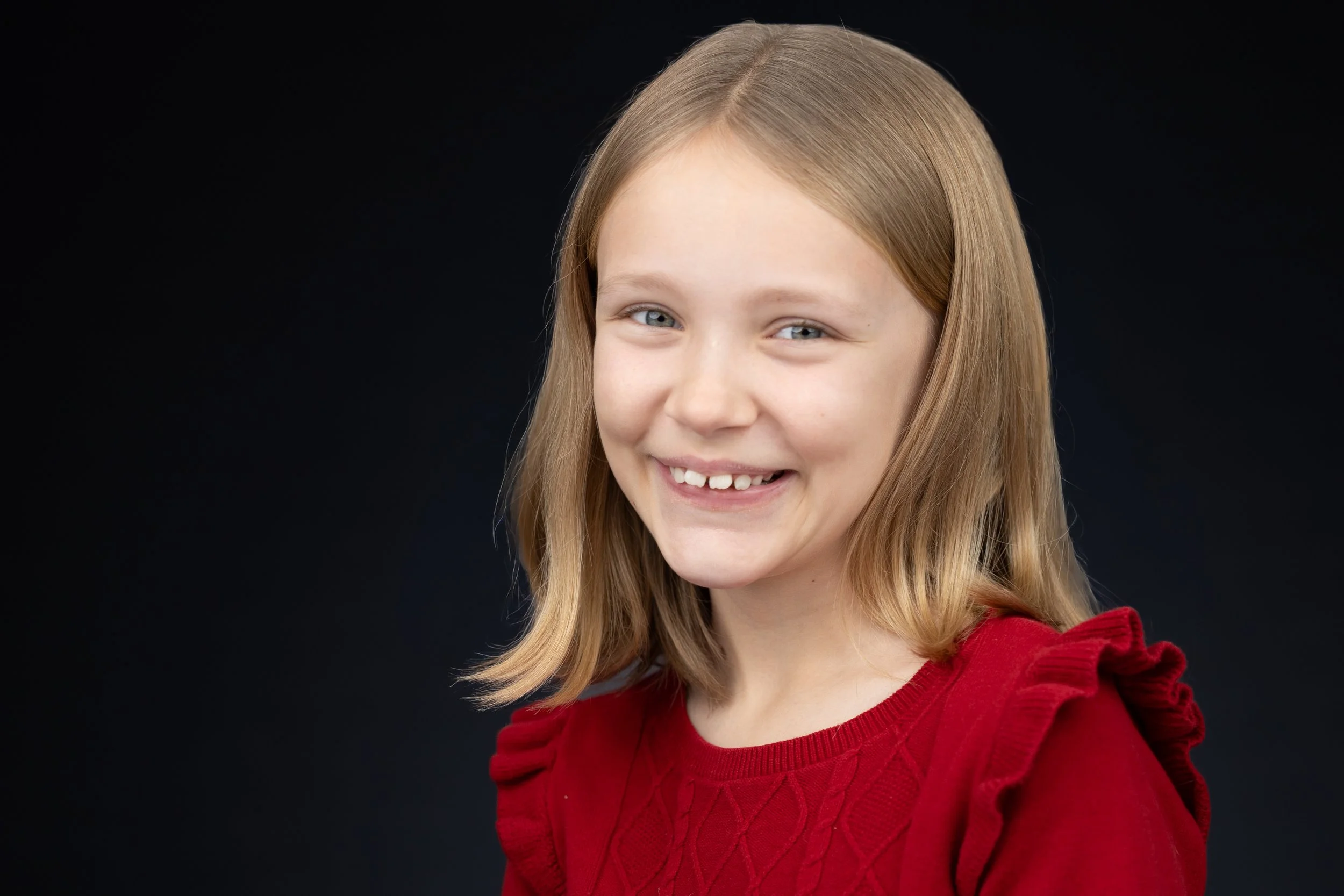 A young girl with blonde hair smiling, wearing a red sweater, against a dark background.