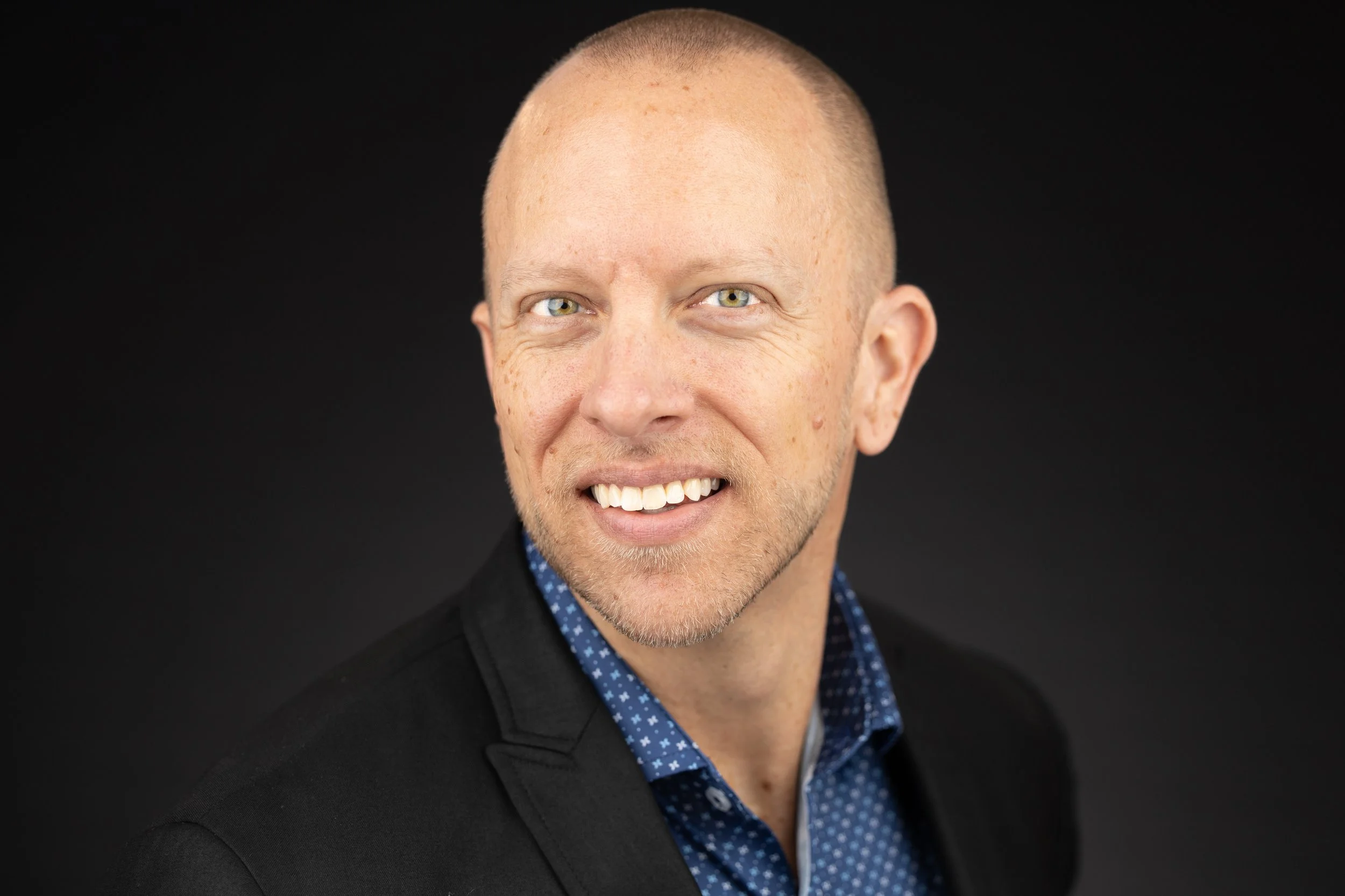 Headshot of a smiling man with light skin, closely shaved head, and blue eyes, wearing a black blazer and blue polka dot shirt, against a black background.