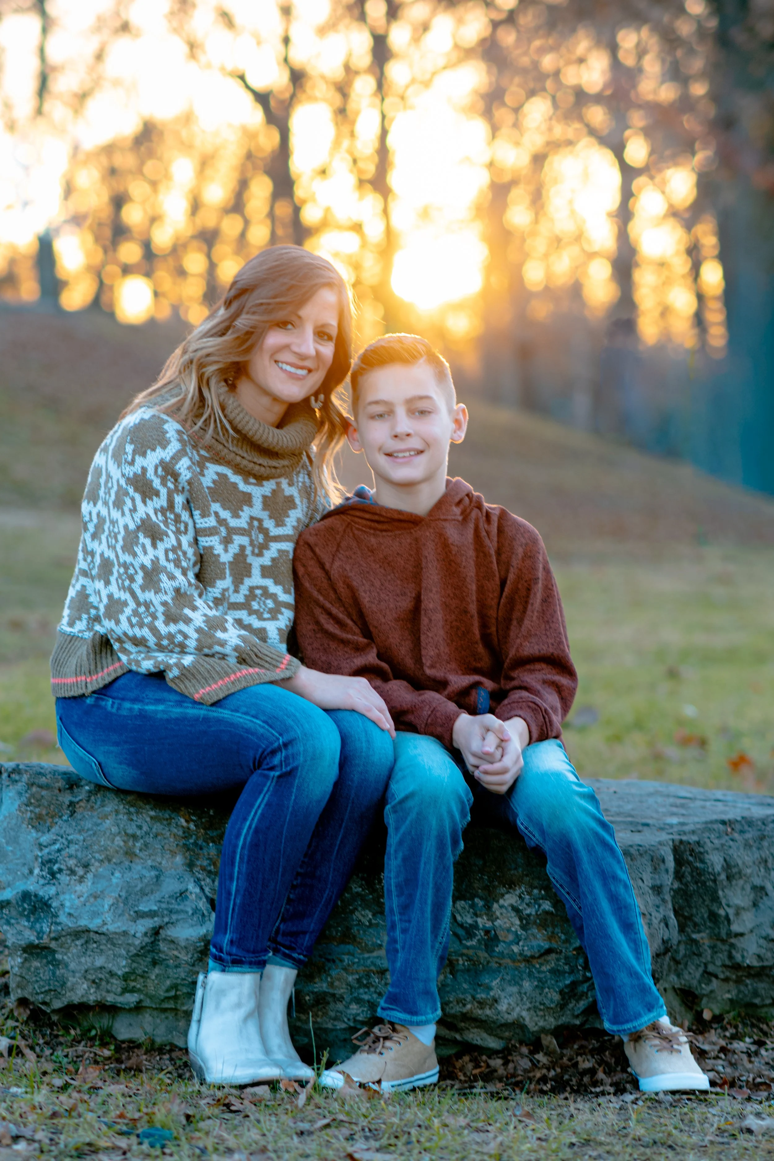 A woman and a young boy sitting on a large stone outdoors during sunset, smiling at the camera. The woman is wearing a patterned sweater and jeans, the boy is in a brown hoodie and jeans, with a background of trees and sunset light.