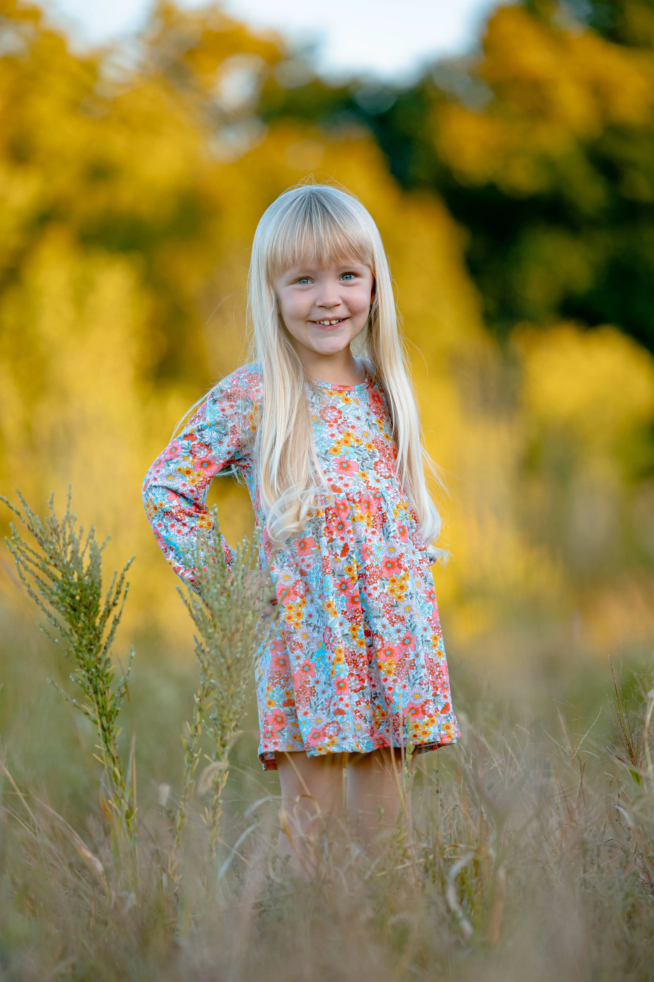 A young girl with long blonde hair wearing a colorful floral dress standing in a grassy field with a background of yellow and green trees during fall.