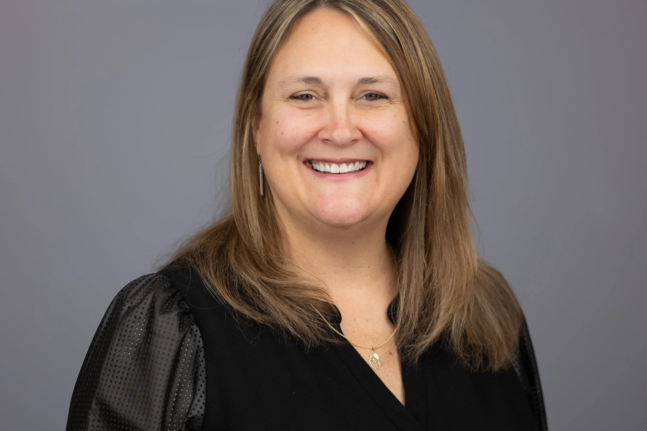 A smiling woman with shoulder-length light brown hair wearing a black top with sheer, dotted sleeves, a gold necklace, and earrings, against a gray background.