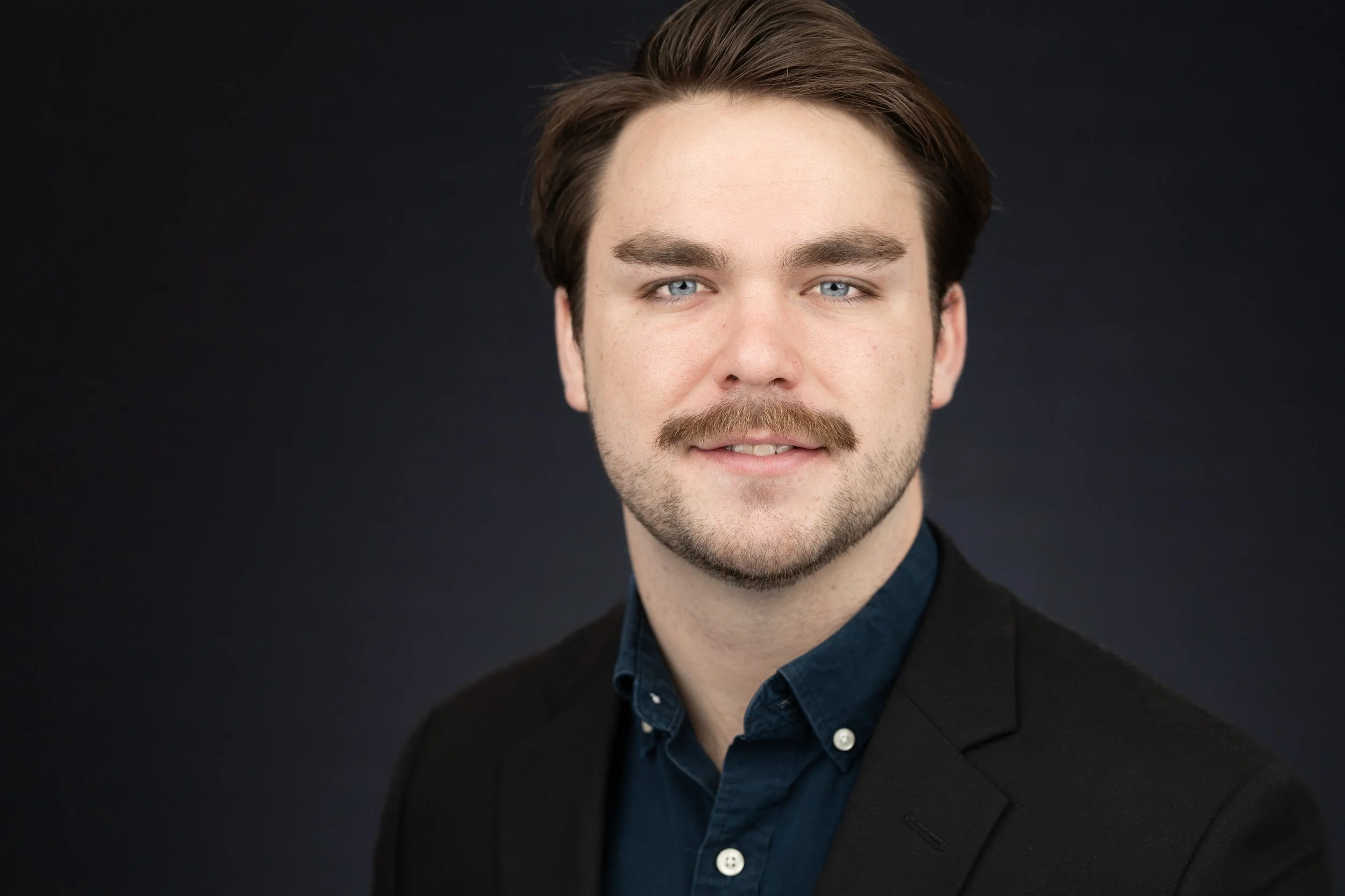 Headshot of a young man with light skin, brown hair, a mustache, and a slight smile, dressed in a dark blazer and a button-up shirt, against a dark background.