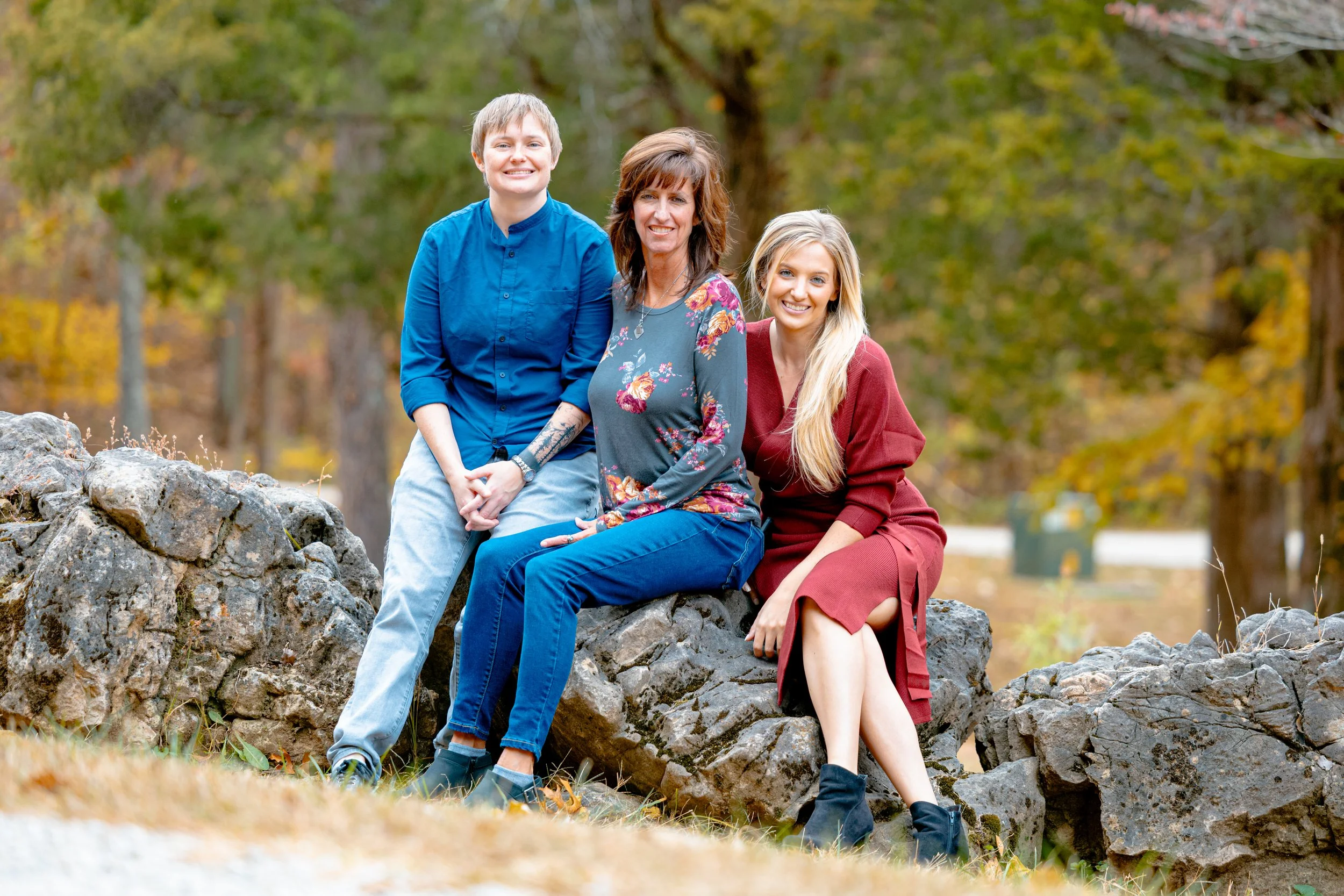 Four women sitting on rocks outdoors in a park with autumn trees in the background, smiling at the camera.