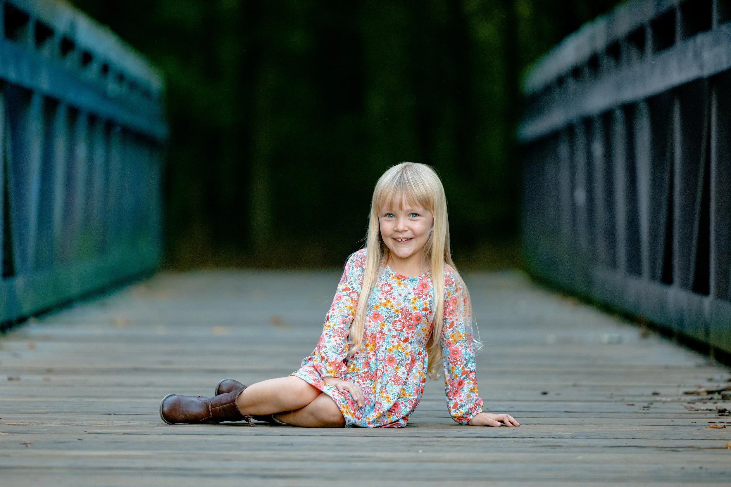 A young girl with long blonde hair in a floral dress and boots sitting on a wooden bridge, smiling at the camera.