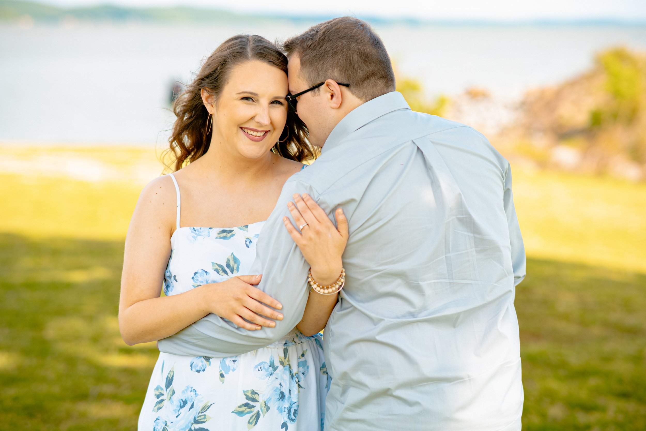 A smiling woman in a floral dress with her arm around a man wearing glasses and a light line shirt, standing outdoors near a lake with trees in the background.