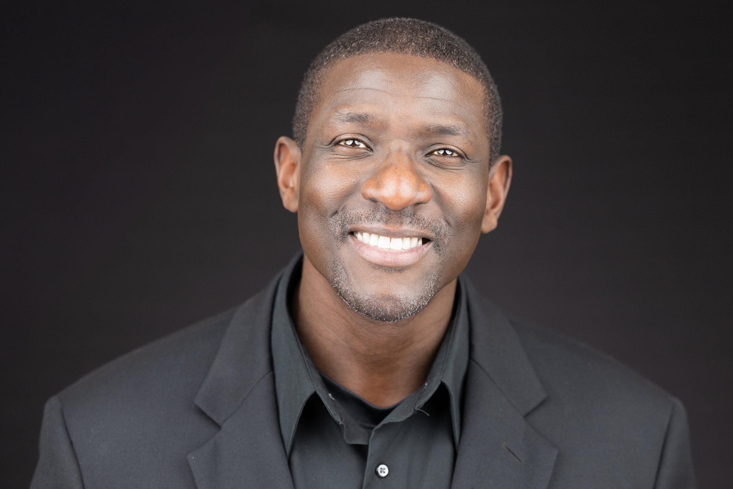 A middle-aged African American man with short hair and a goatee, smiling, dressed in a dark blazer and shirt, against a black background.