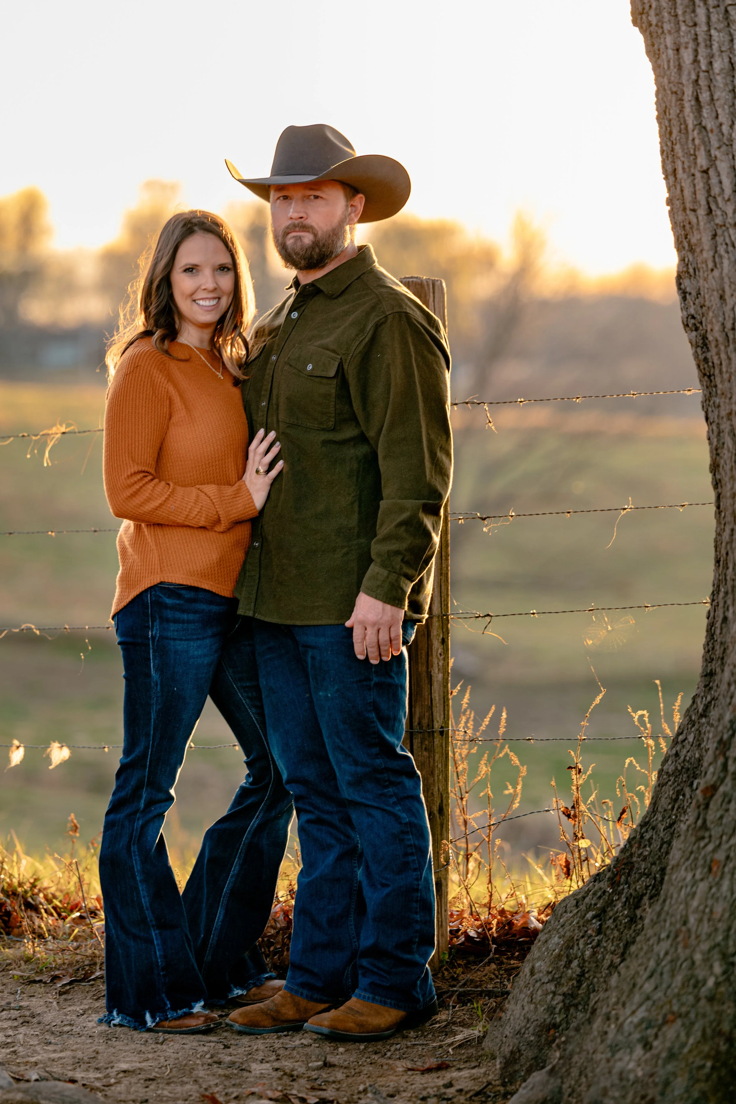 A man and woman standing outdoors near a tree and a barbed wire fence, with a sunset background. The woman wears a rust-colored sweater and jeans, and the man wears a cowboy hat, an olive green shirt, and jeans.