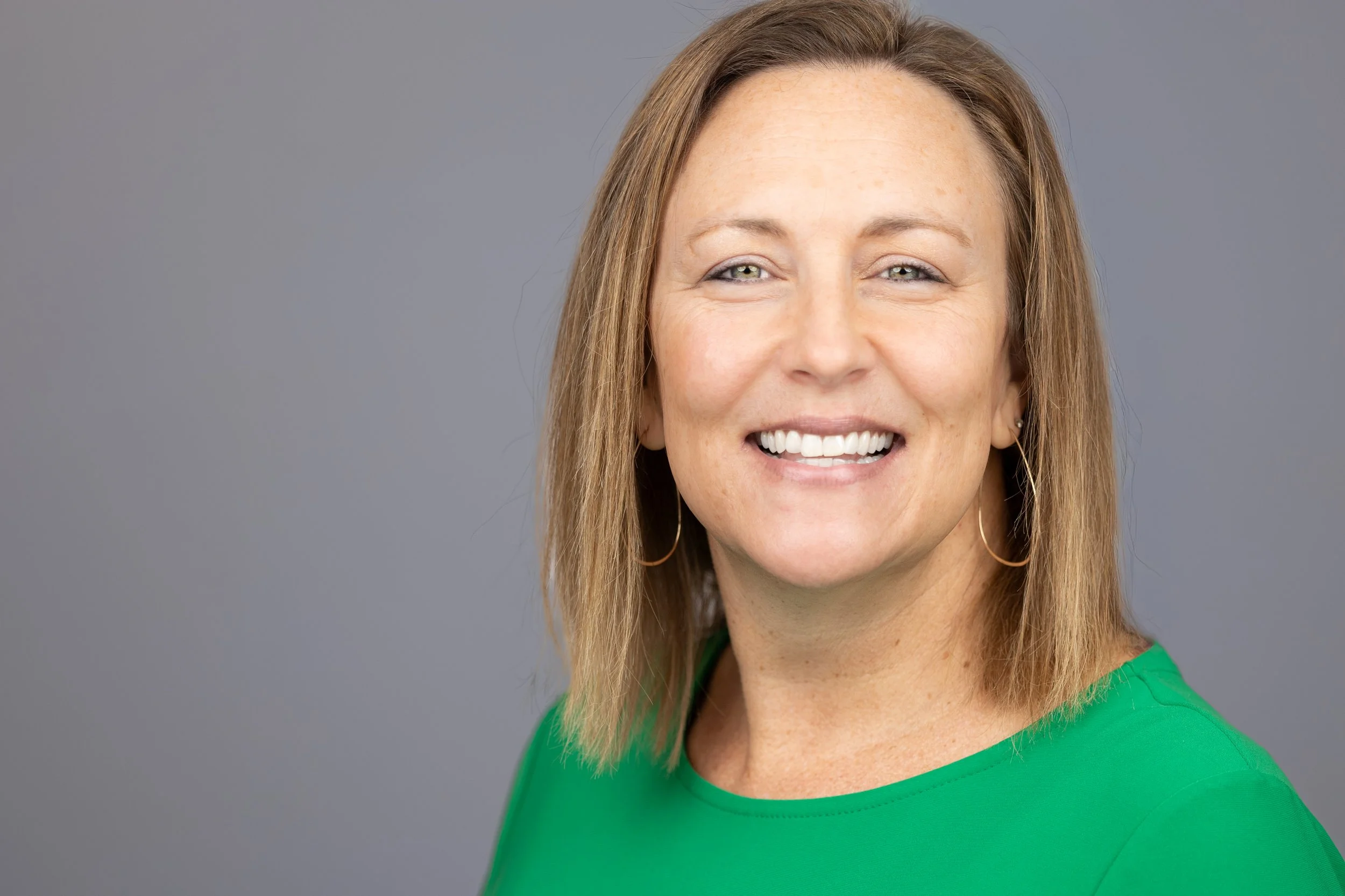 A woman with shoulder-length light brown hair smiling, wearing a green top and hoop earrings, against a plain gray background.