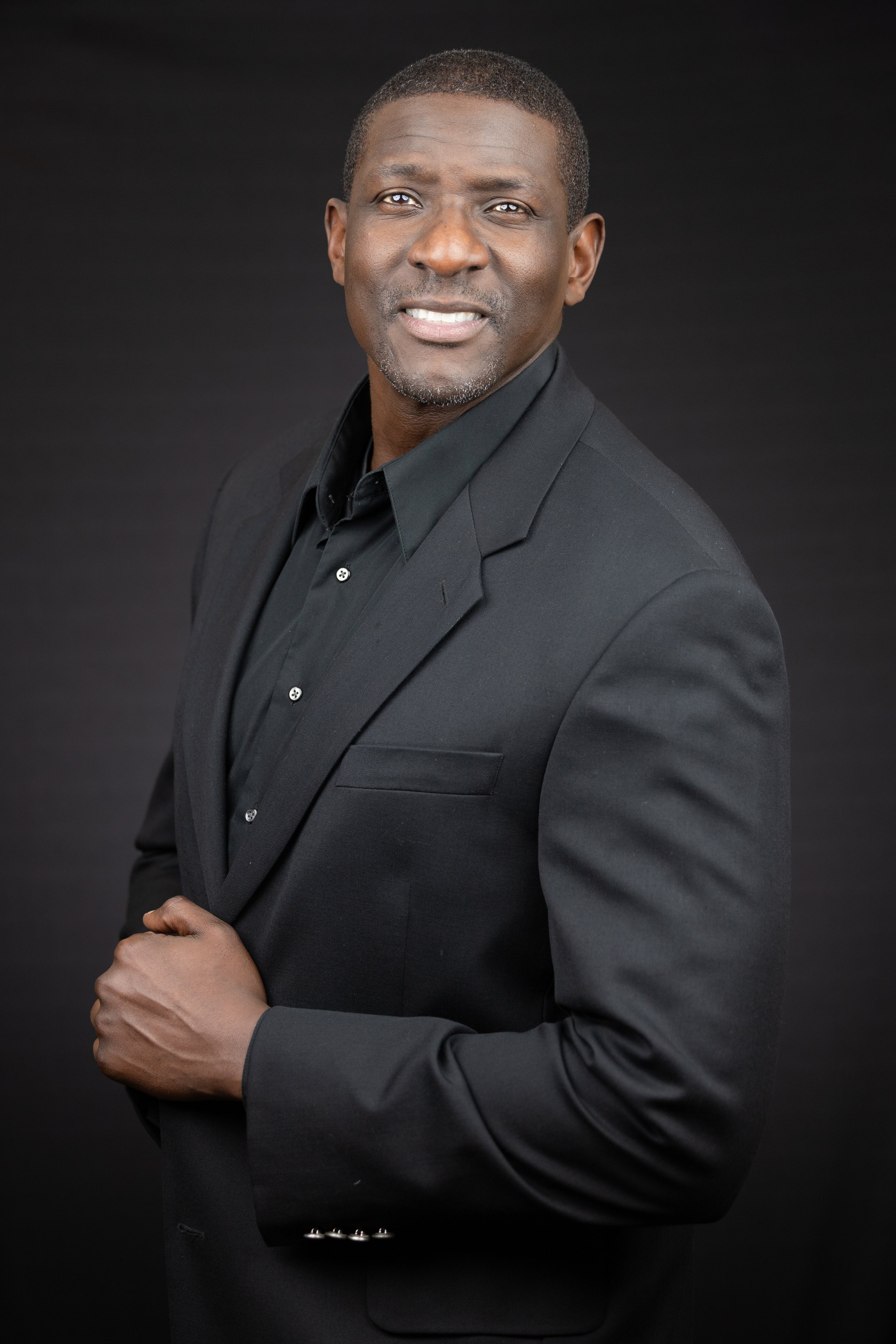 A confident African American man in a black suit, black shirt, and no tie, standing against a black background.