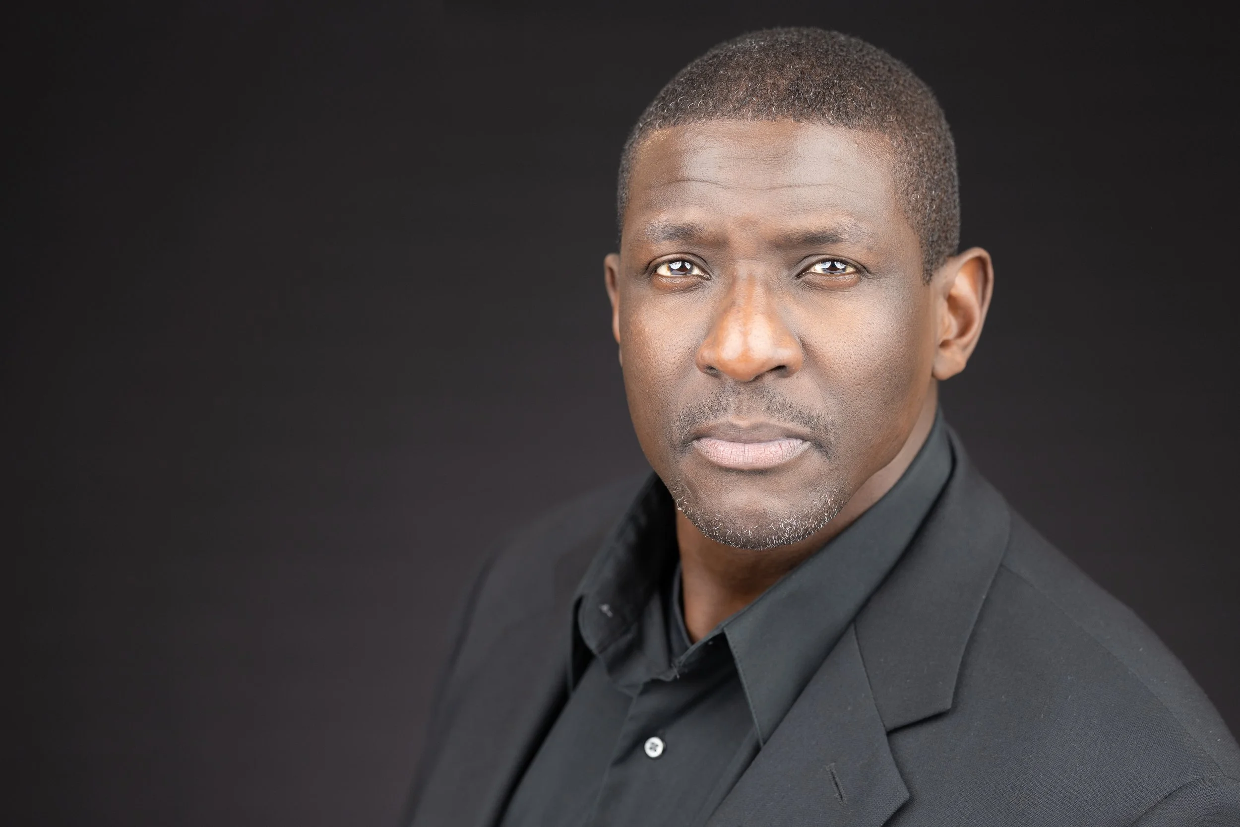Professional portrait of a middle-aged African American man with short hair, wearing a black suit and shirt, looking directly at the camera against a black background.