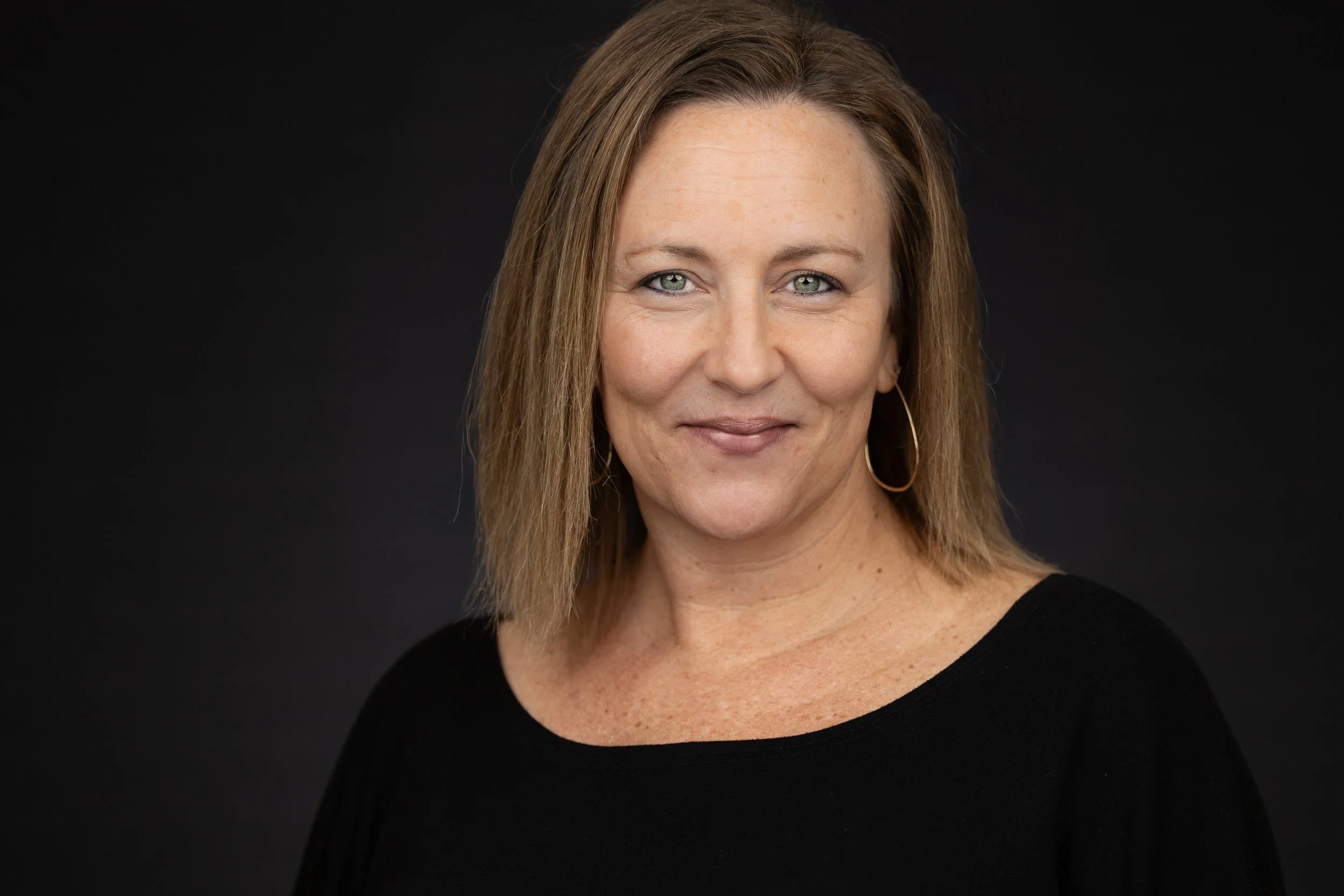 A middle-aged woman with shoulder-length light brown hair, blue eyes, and gold hoop earrings, wearing a black top, smiling slightly against a solid black background.