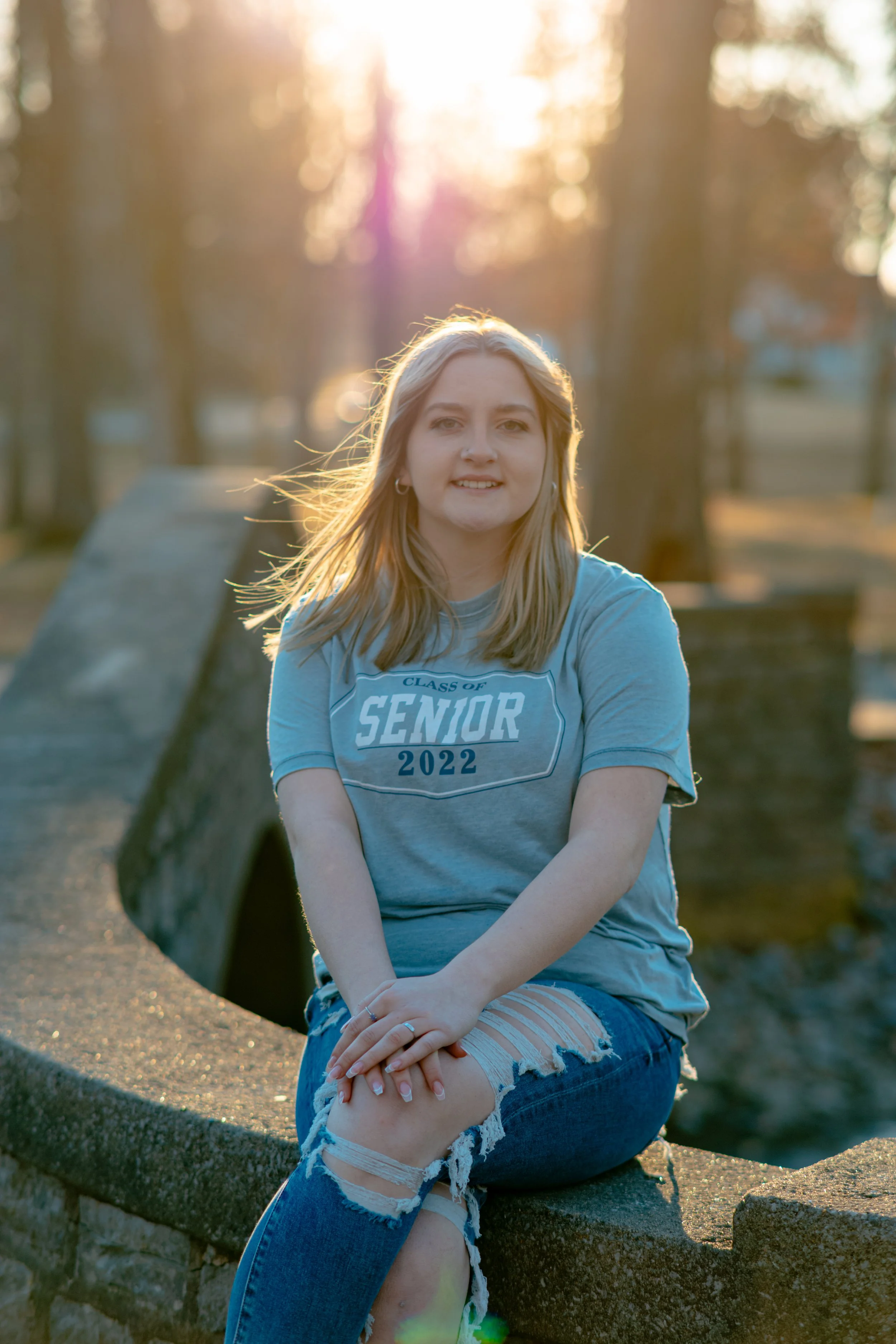 Young woman with blonde hair, sitting on a stone bench outdoors during sunset, wearing a gray 'Class of Senior 2022' T-shirt and ripped jeans.