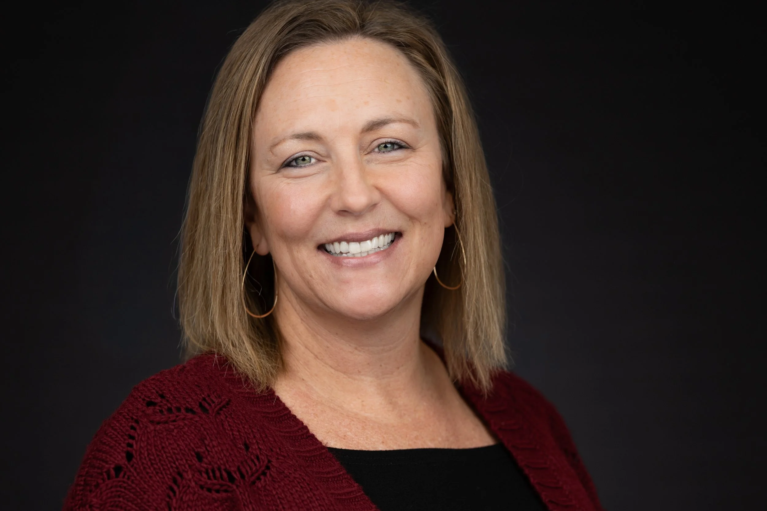 A woman with shoulder-length light brown hair, smiling, wearing a black top and a red cardigan, against a dark background.
