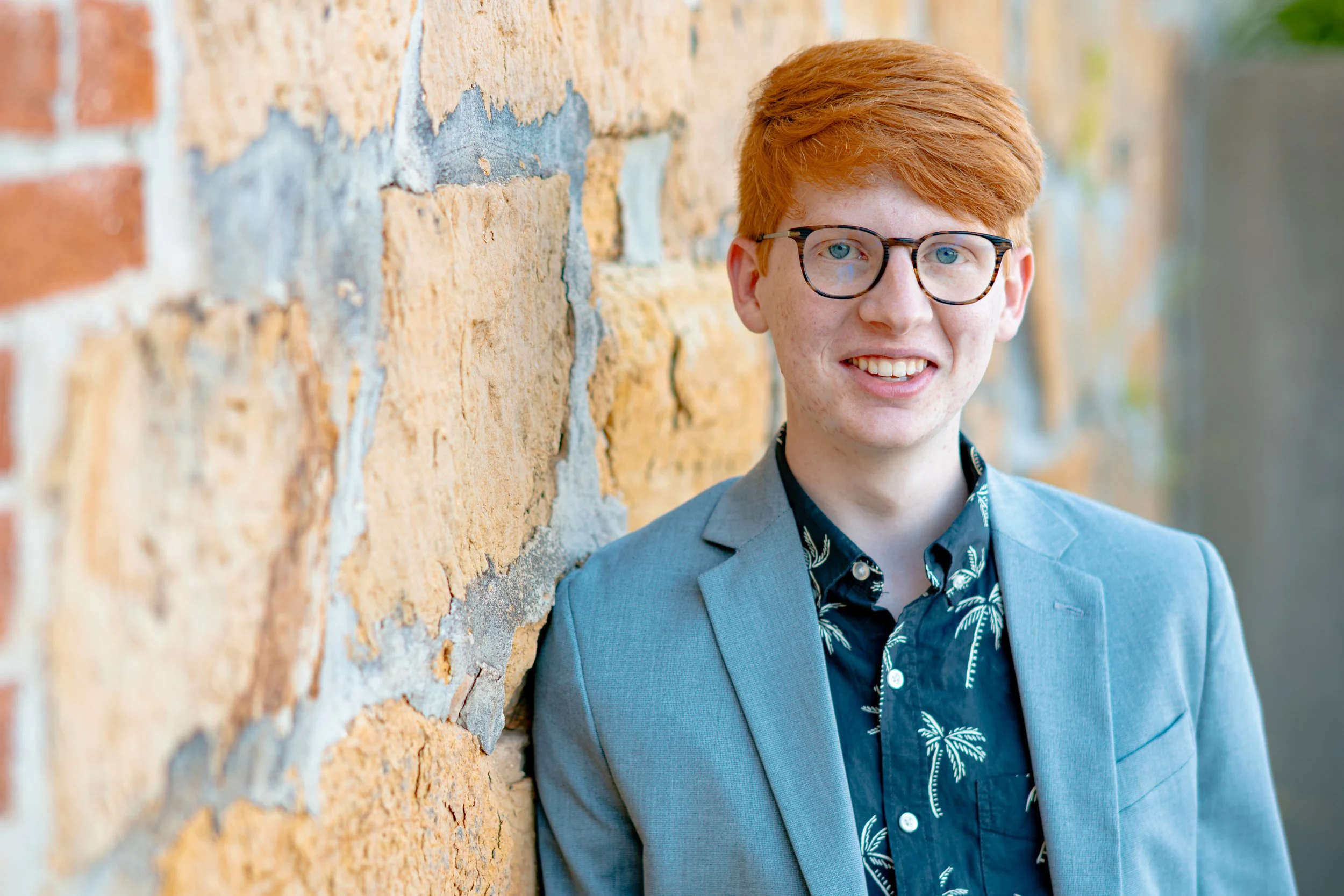 Young man with red hair, glasses, and a blue suit jacket, smiling, standing outdoors next to a brick wall.