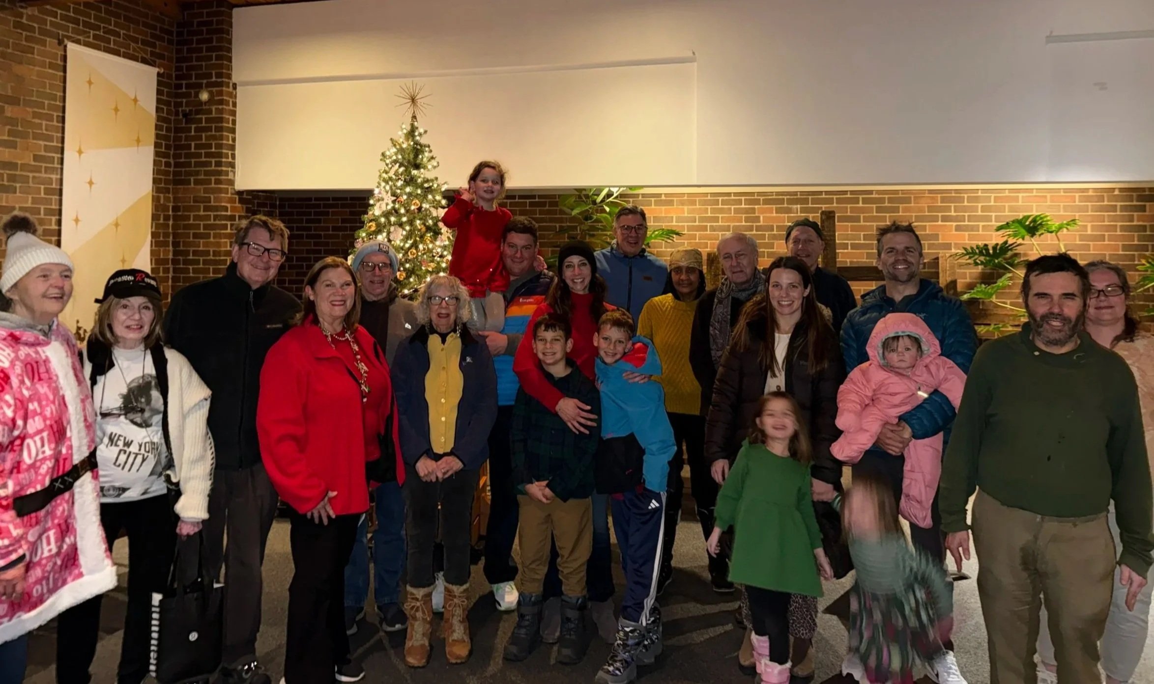 A group of adults and children posing indoors in front of a decorated Christmas tree at Church of the Tree Crosses, with brick walls and indoor plants visible.
