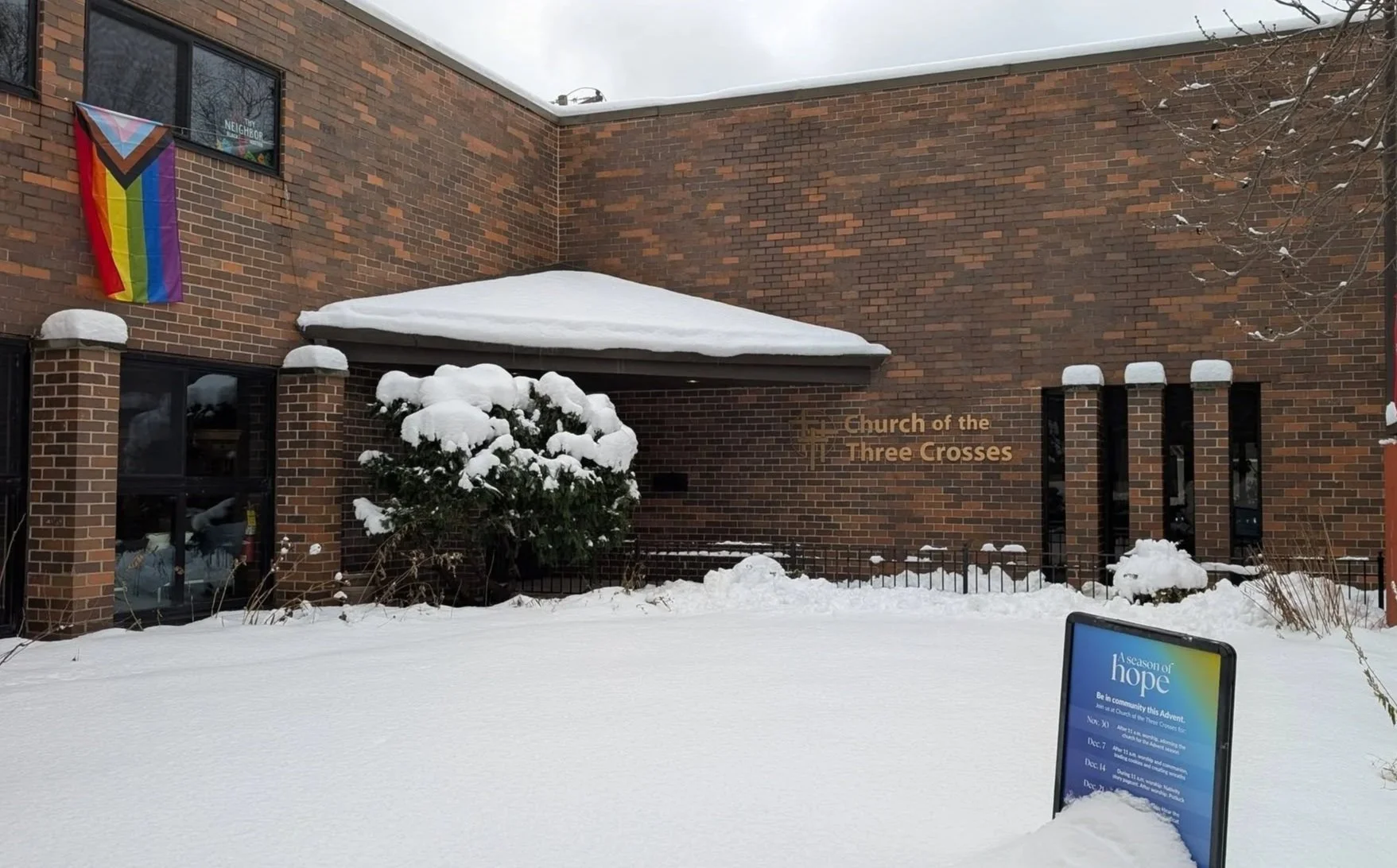 Snow-covered exterior of the Church of the Three Crosses with rainbow pride flag hanging beside a window, snow-laden shrub, and a sign displaying information about a community event.