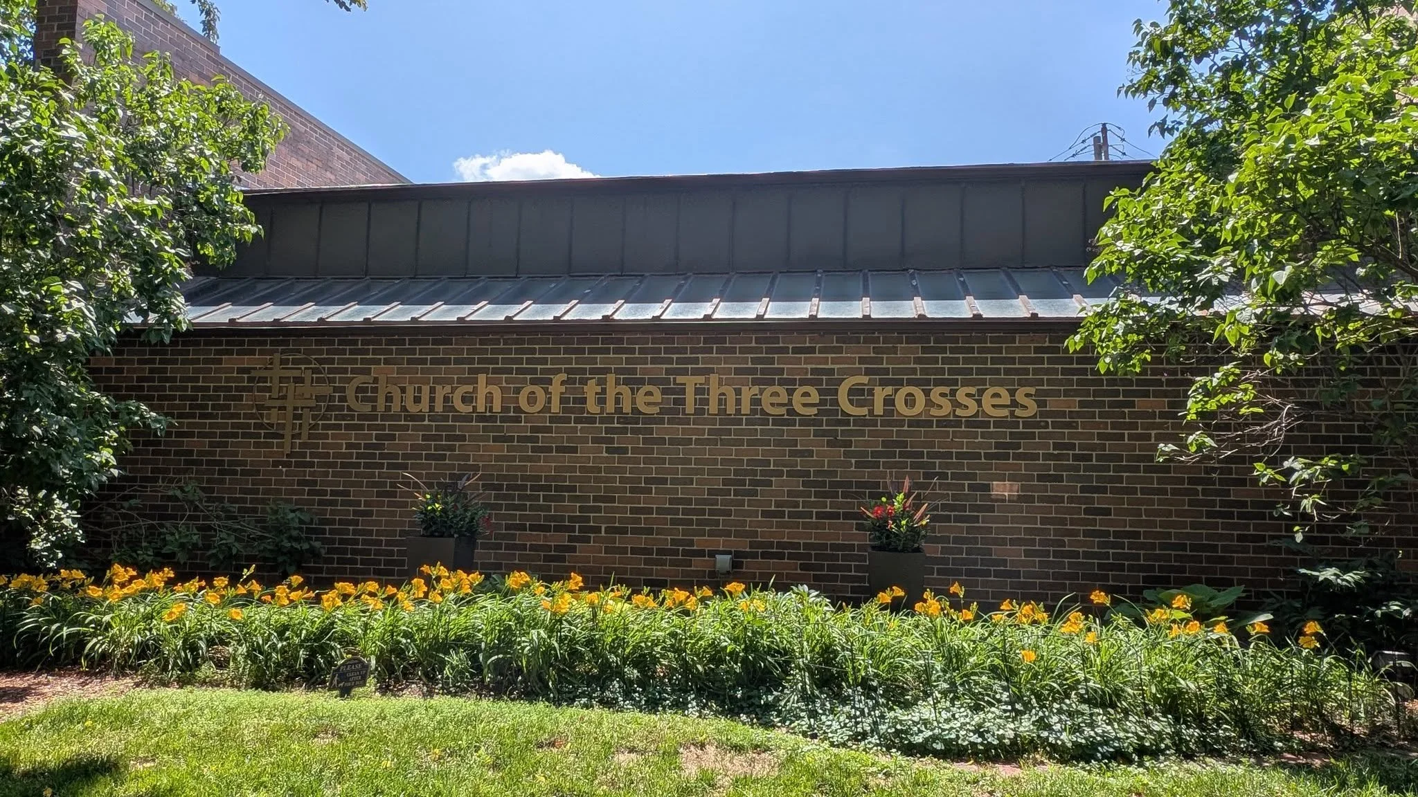 Brick building with gold lettering that says 'Church of the Three Crosses' and a cross logo, surrounded by green trees and yellow flowers, under a blue sky with some clouds in the Old Town Triangle part of Chicago..