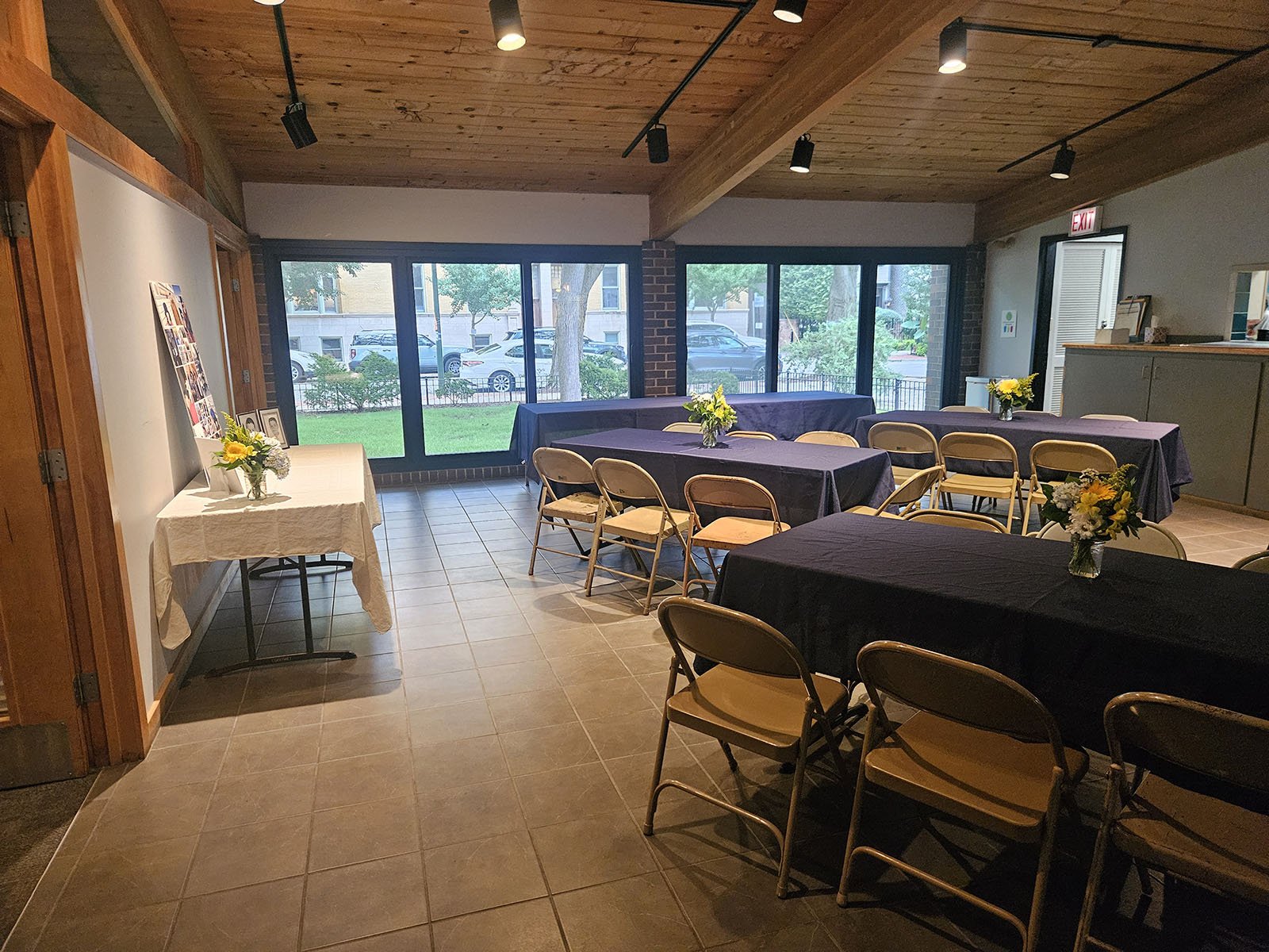 Church of the Three Crosses' indoor event space with tables covered in dark tablecloths, decorated with vases of yellow and white flowers, and chairs arranged around the tables. Large windows show an outside view of trees and a lawn for events.