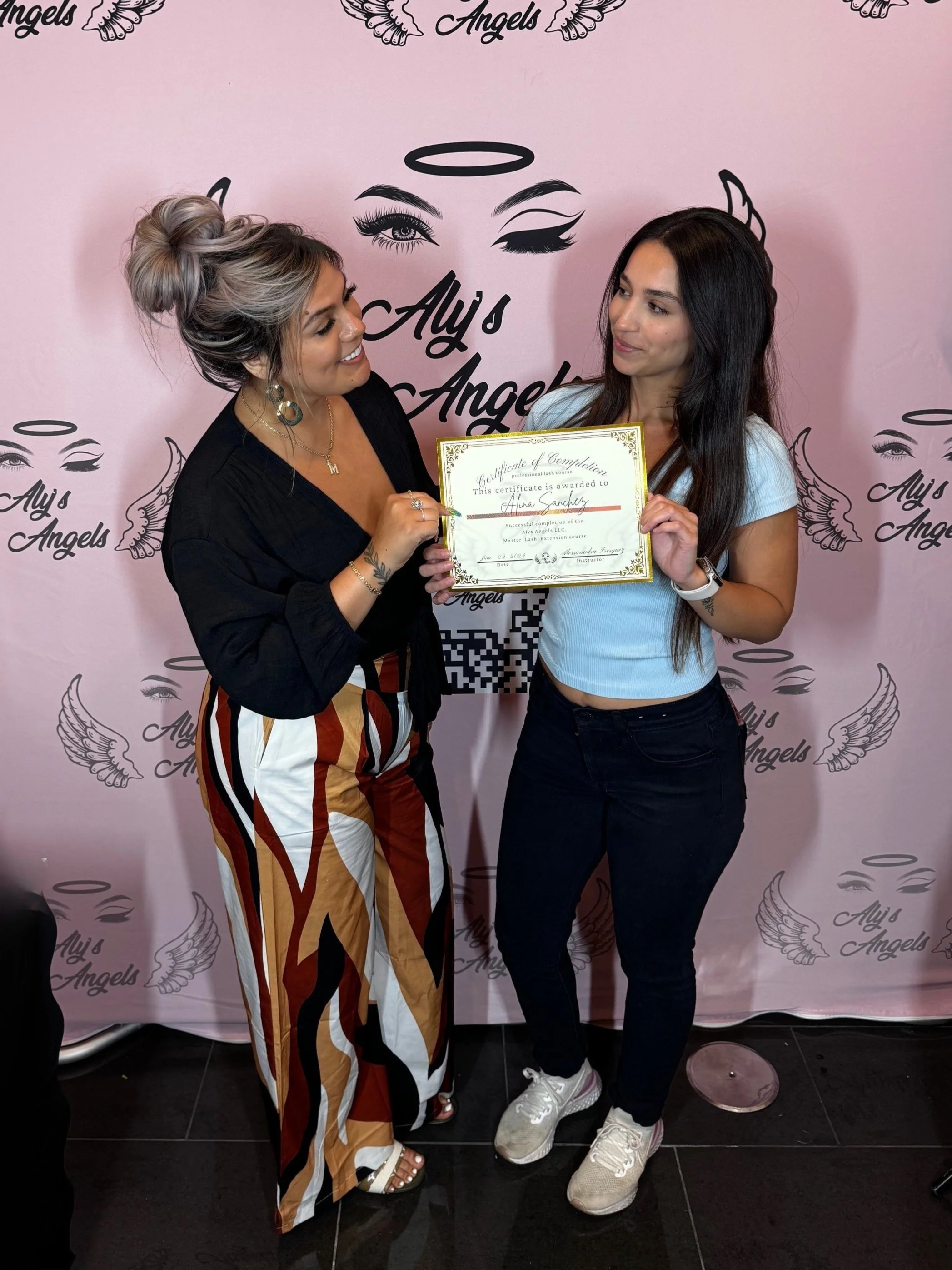 Two women standing together, one is presenting a certificate to the other. The background has pink and black text and graphics, including the words "Aly's Angels" and angel wing illustrations.