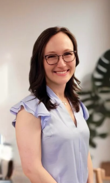 A smiling woman with glasses and dark hair, wearing a light purple blouse with ruffled sleeves, standing indoors with a blurred plant in the background.