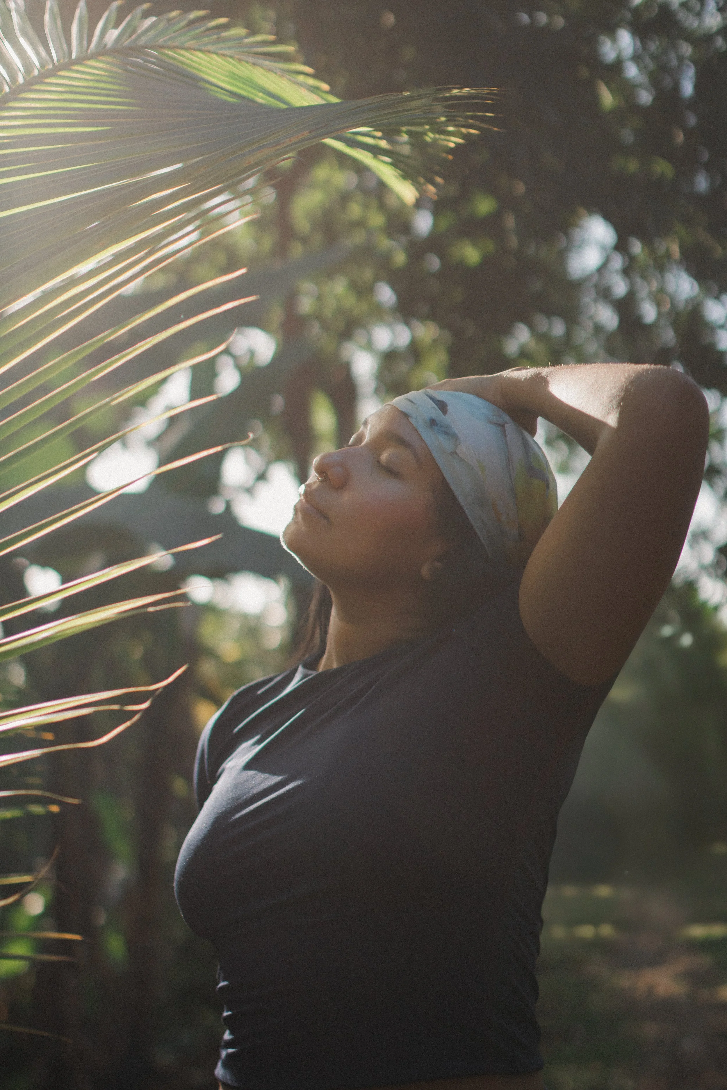 Woman with closed eyes, wearing a headscarf, outdoors in a sunny, leafy environment, appearing peaceful and relaxed.