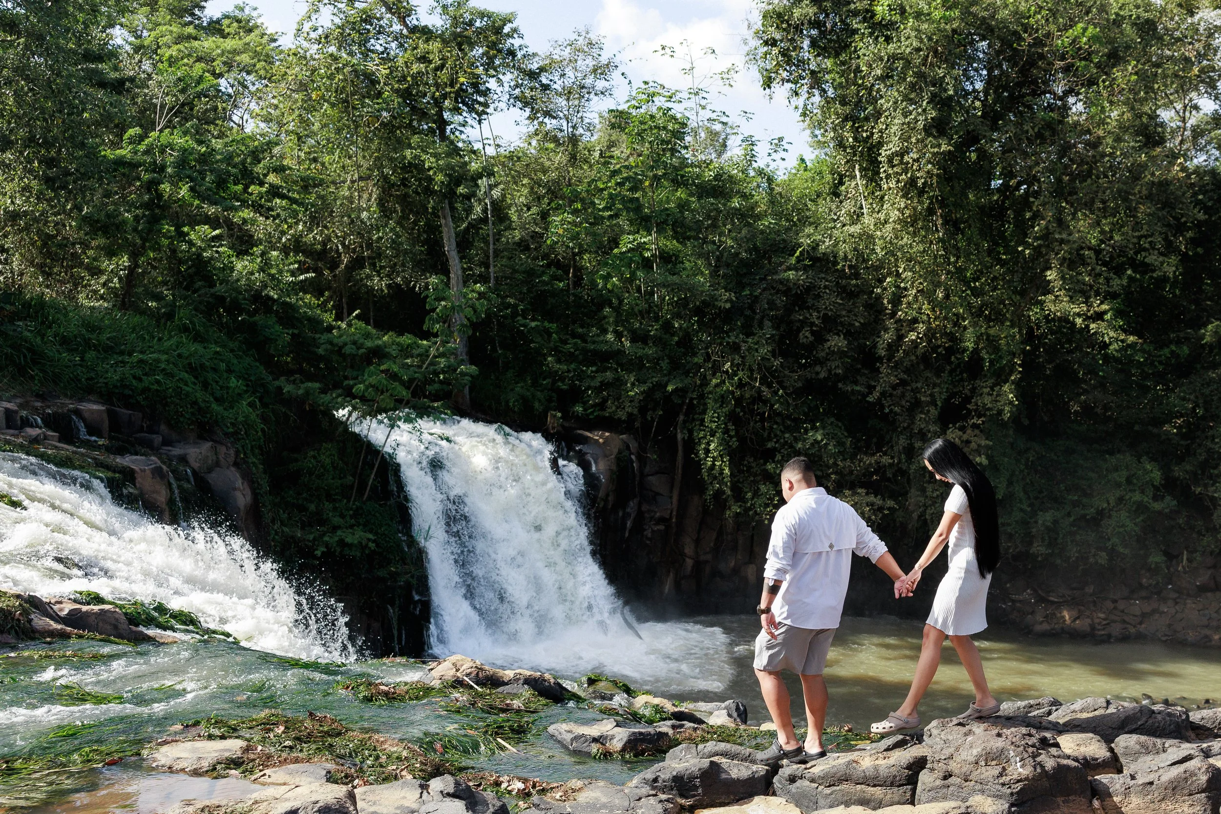 A couple holding hands on rocks near a waterfall in a lush green forest.