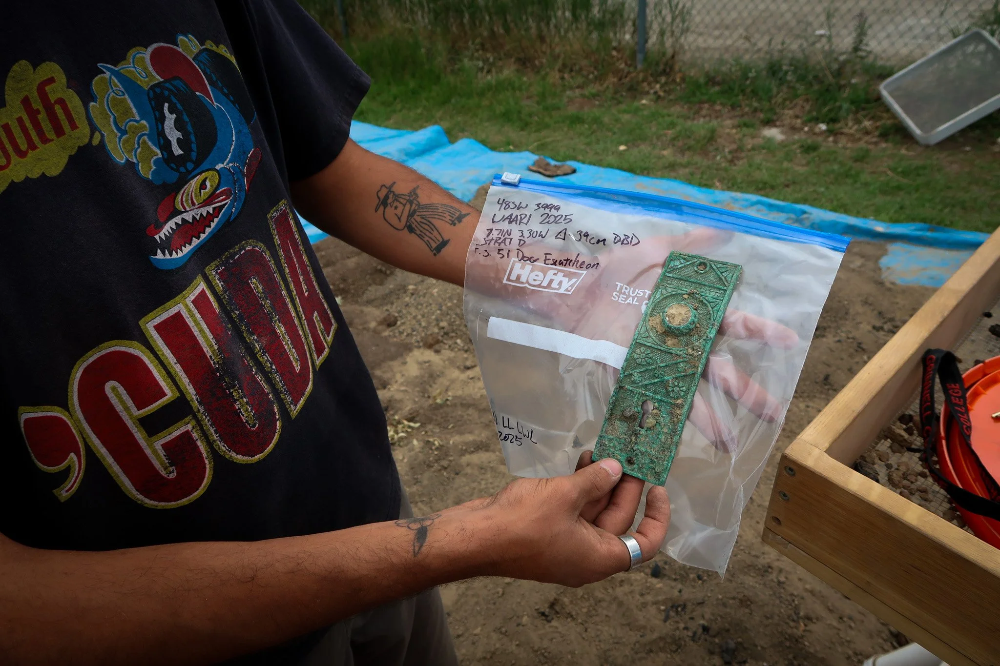 Person holding a plastic bag containing a metal object outdoors. The person has tattoos on their arm and is wearing a black t-shirt with colorful graphics.