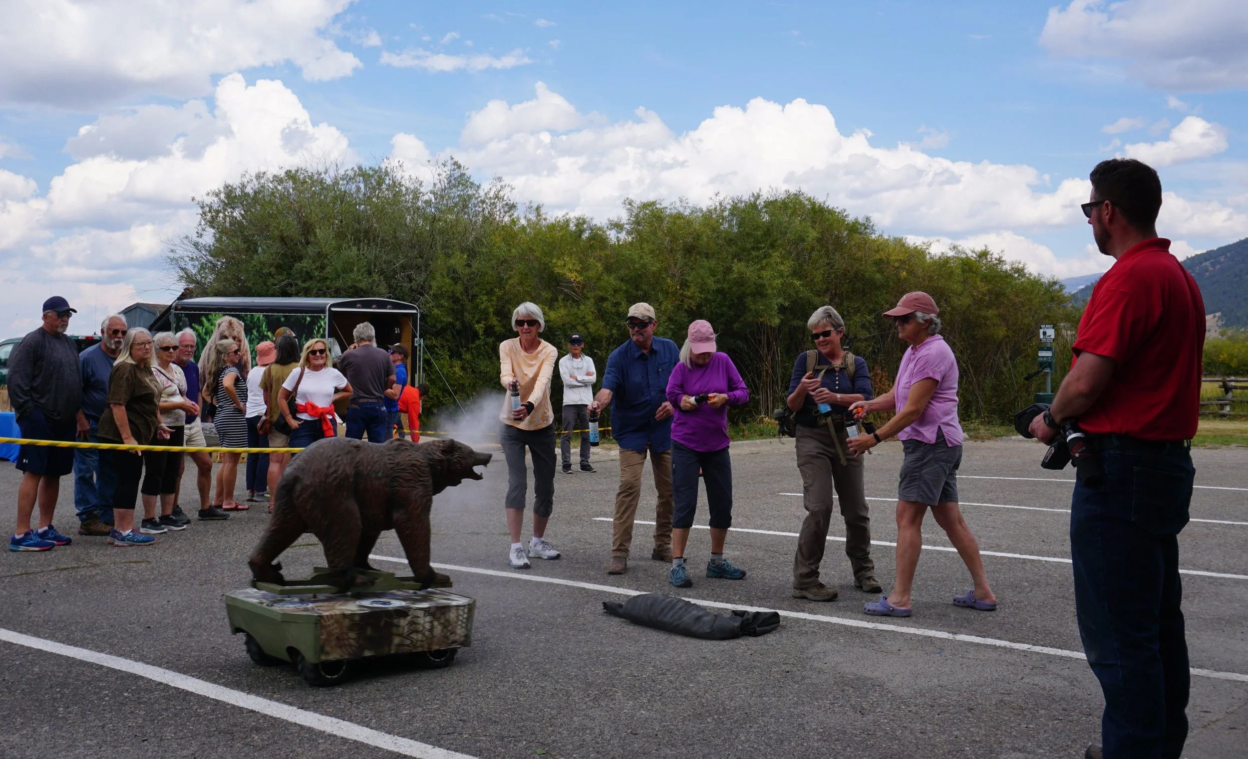 Group of people gathered outdoors during daytime, with some taking photos or videos of a bear. The scene includes children and adults, some wearing hats and sunglasses, with a bear walking near them on a paved area. There are trees and mountains in the background under a partly cloudy sky.