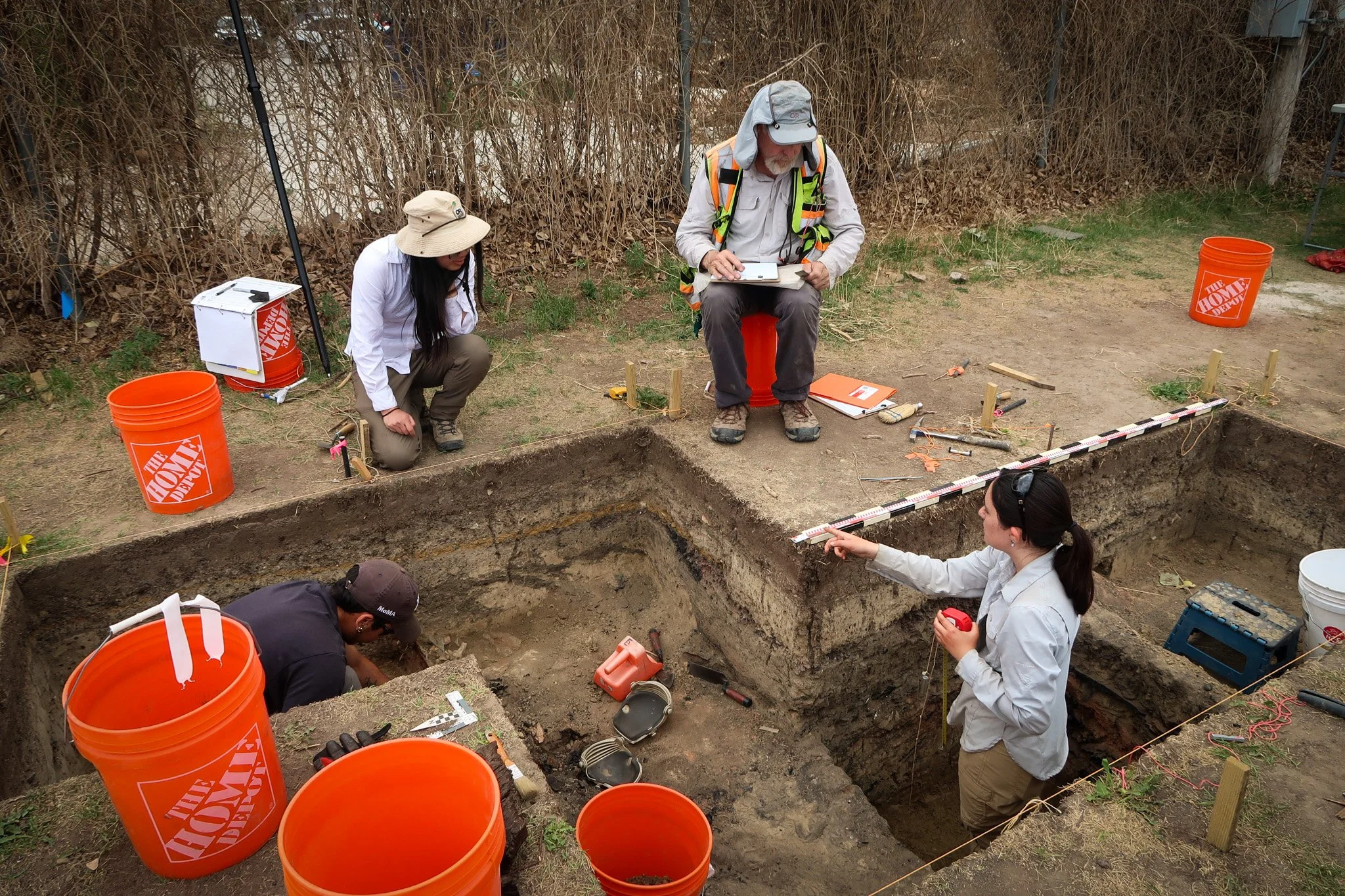 Archaeologists excavating an archaeological site with tools and measuring equipment, with orange buckets nearby, under an outdoor setting.