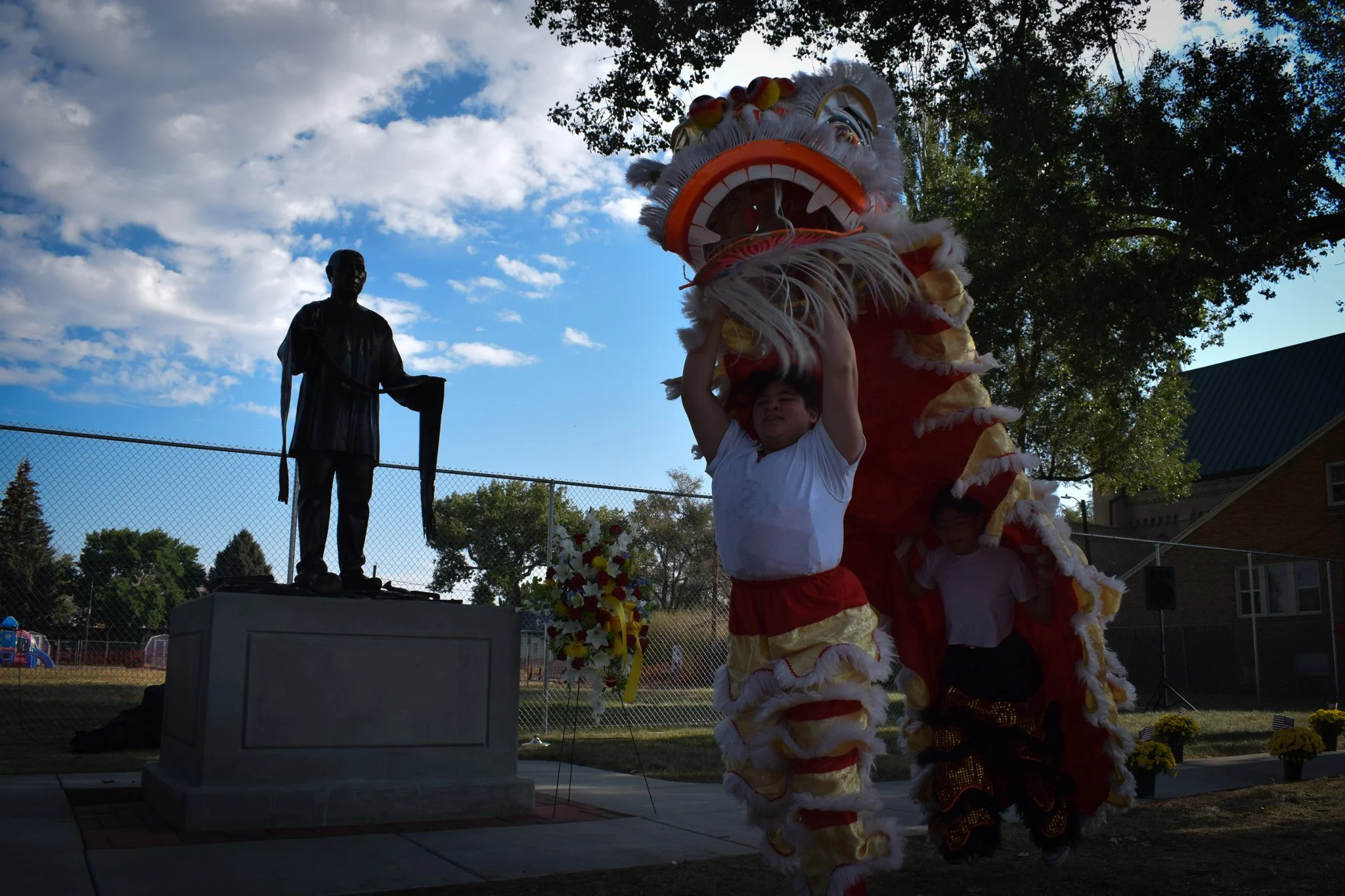 Children participating in a cultural celebration, with a traditional Chinese lion dance costume, and a statue of a man on a pedestal in the background, set outdoors during daytime.