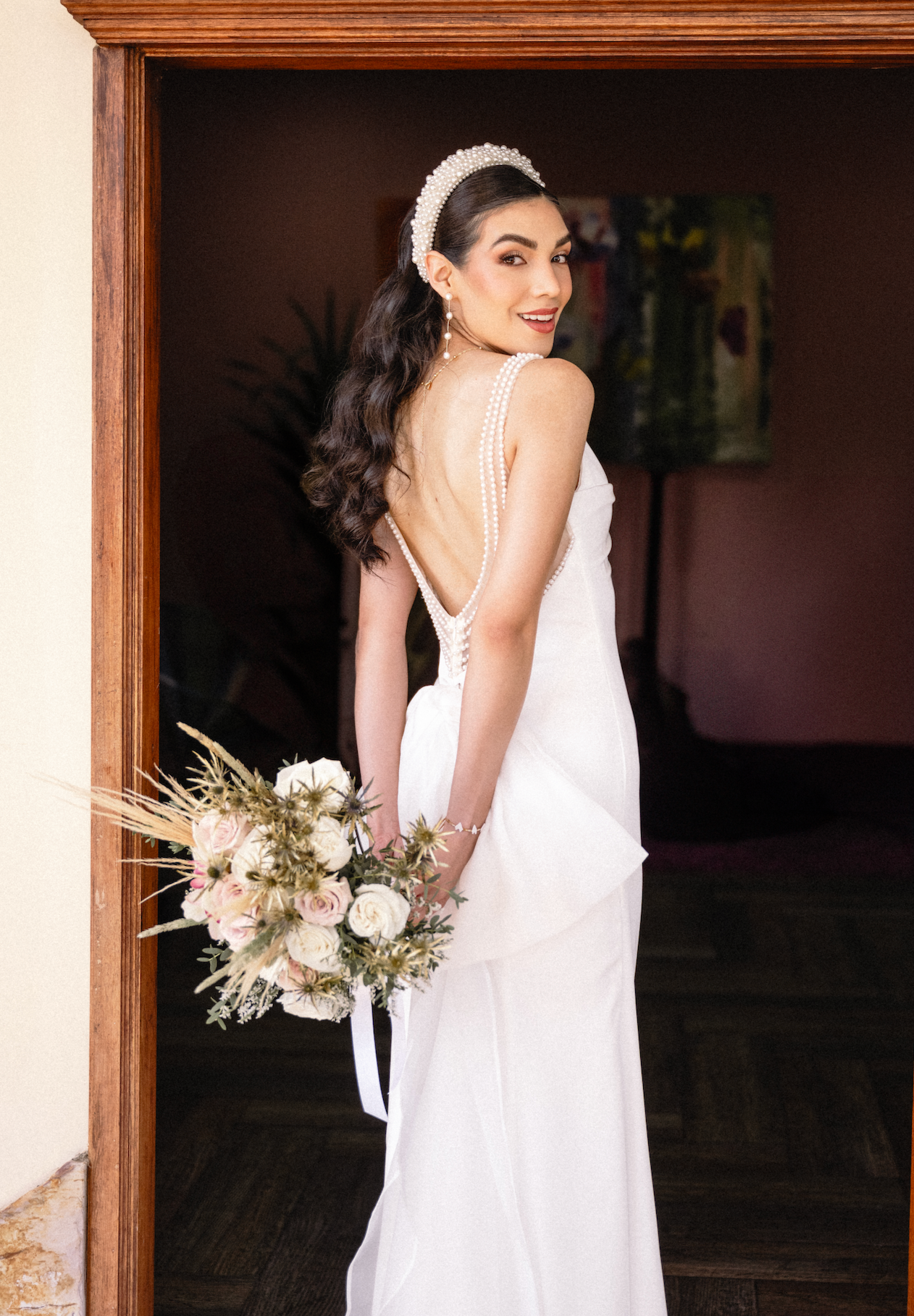 A woman in a white wedding dress with pearl details, holding a bouquet of flowers, standing in a doorway and looking over her shoulder.