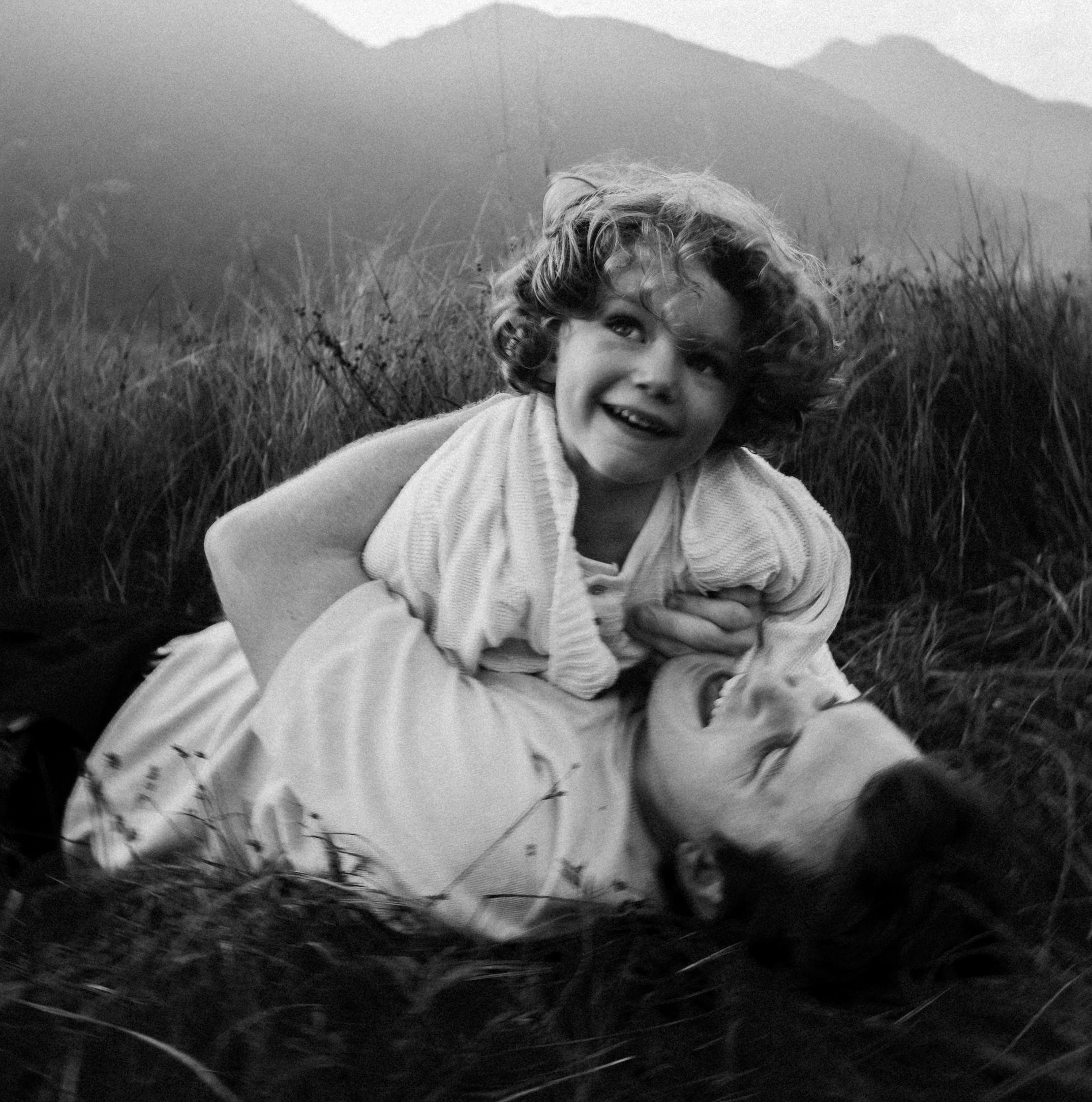 A young girl and an adult woman lying in tall grass outdoors, smiling and playing together, with mountains in the background.