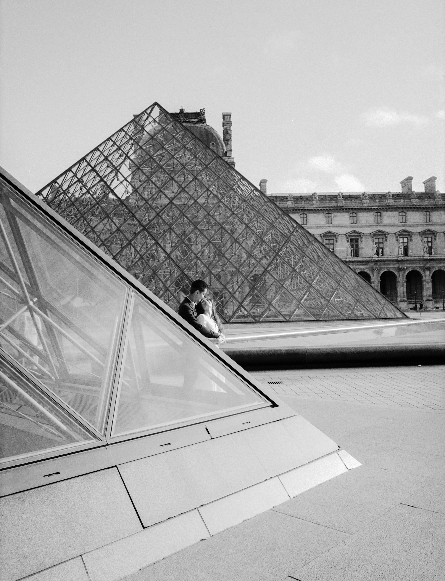 A black and white photo of a couple sitting on a ledge in front of the glass pyramid at the Louvre Museum in Paris, France.
