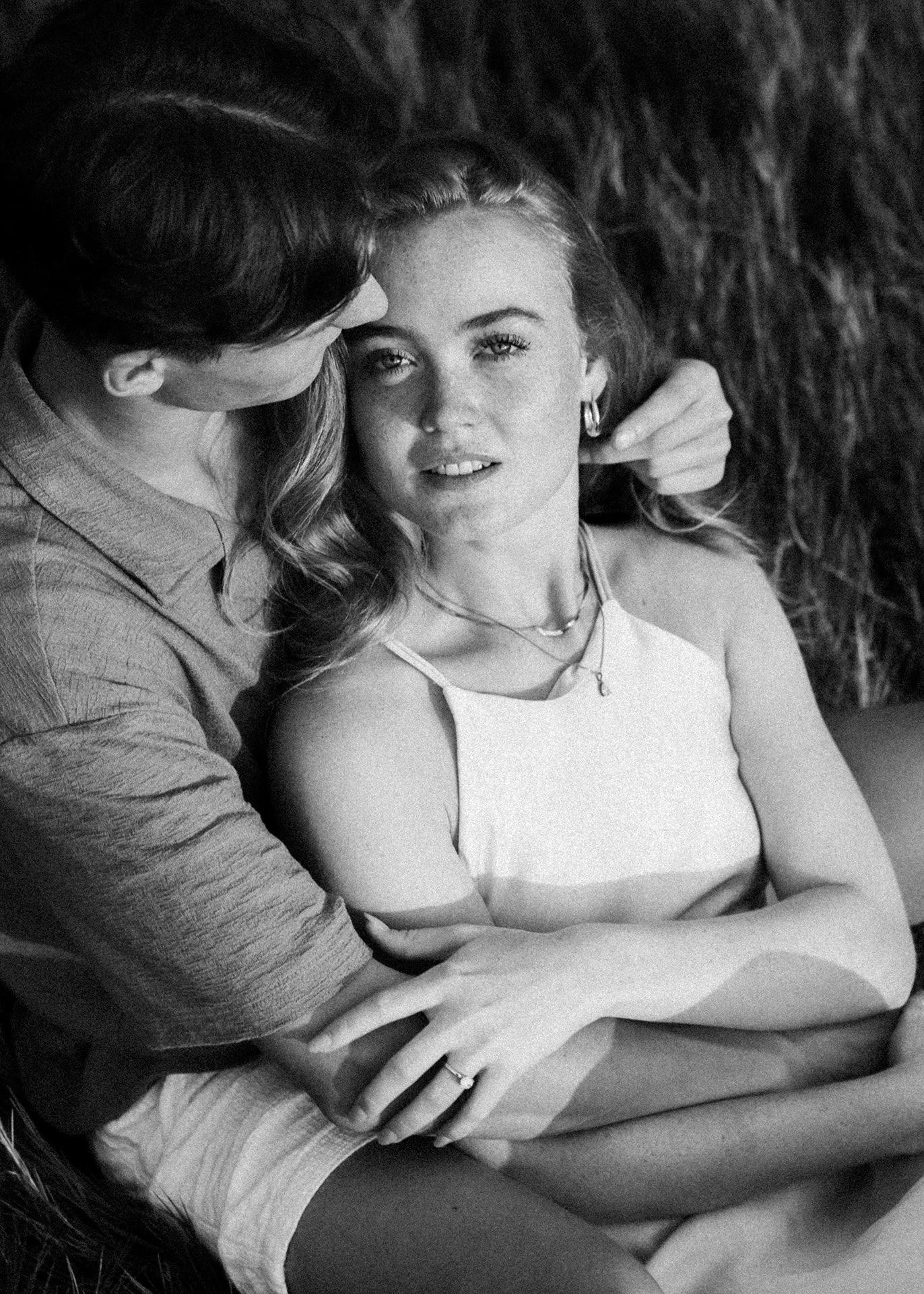 A black and white photograph of a man and woman embracing. The man is kissing the woman on the forehead while she is looking directly at the camera. She has long hair, is wearing earrings and a necklace, and a sleeveless top. The woman's arms are crossed over her lap.